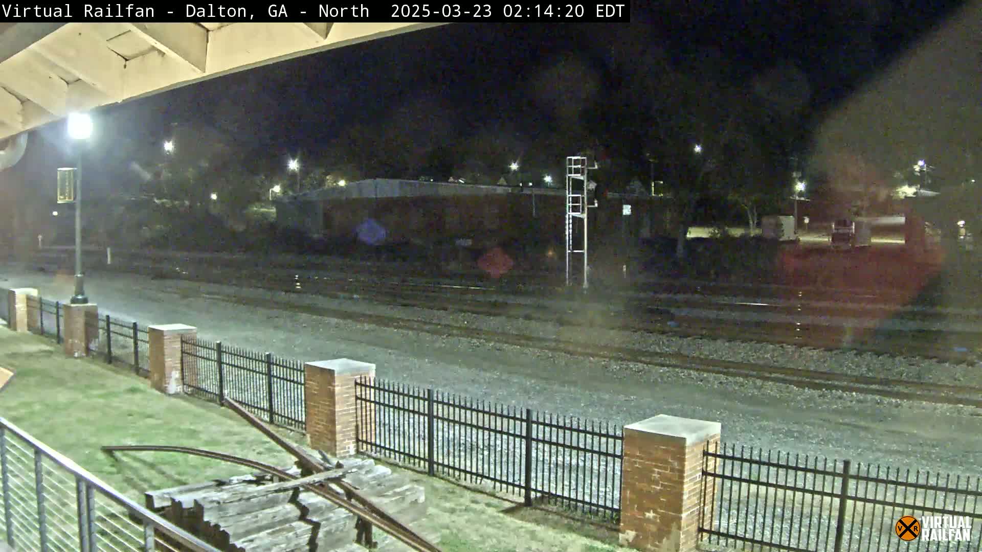 A nighttime view of railroad tracks behind a brick fence and grassy area, under a clear sky.