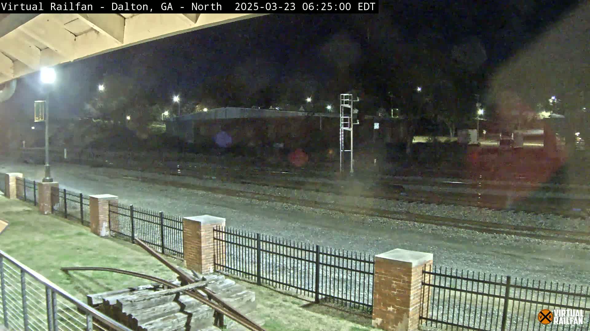 A nighttime view of railroad tracks, bordered by a brick fence and grassy area, with a few distant lights visible.