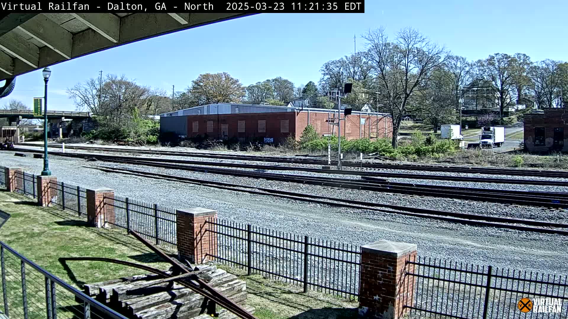 Multiple sets of train tracks extend into the distance under a mostly clear sky, bordered by a brick fence, a grassy area, and buildings in the background.