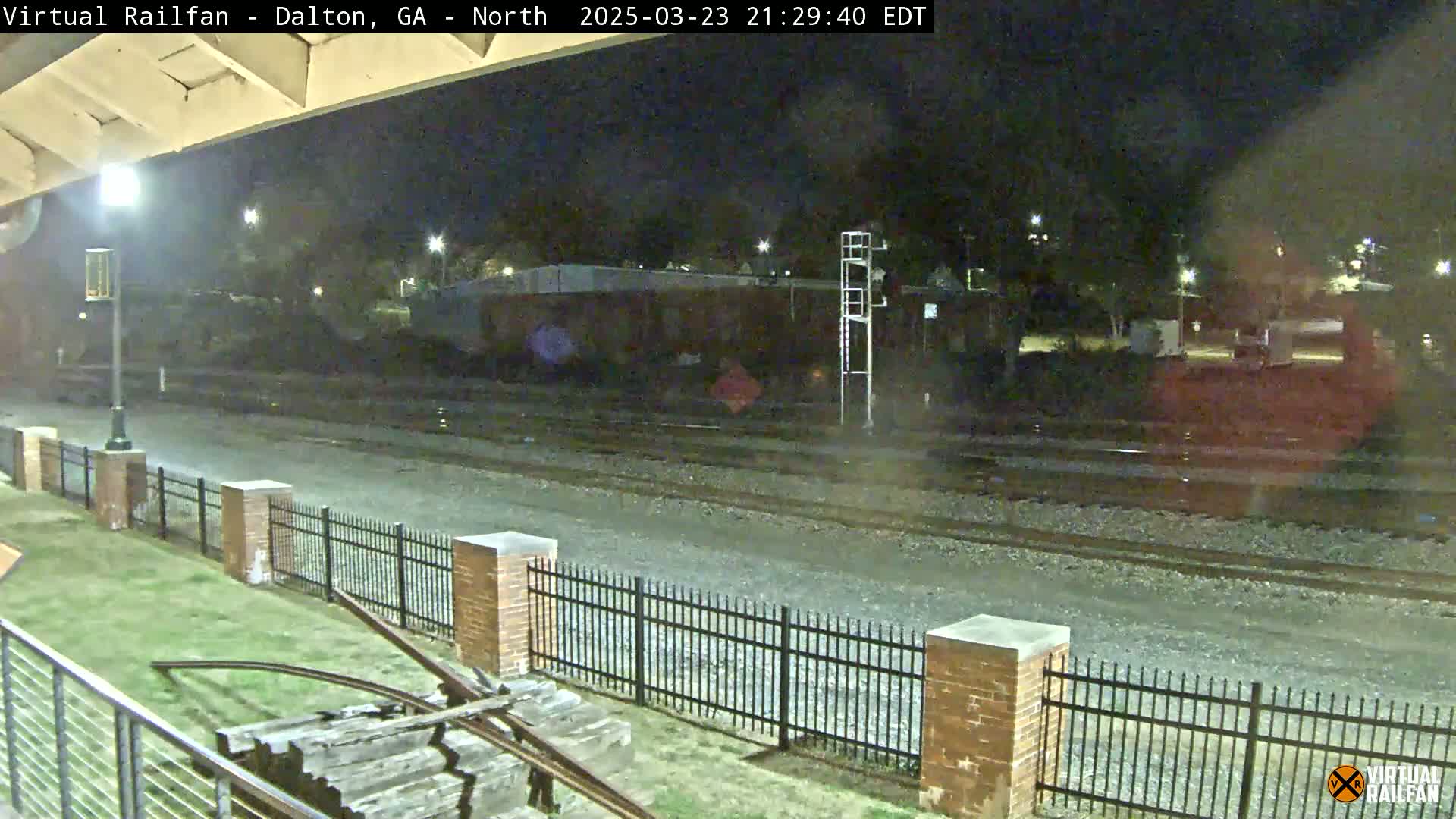 A nighttime view of railroad tracks behind a brick fence, with a grassy area and some old wooden railway equipment in the foreground.