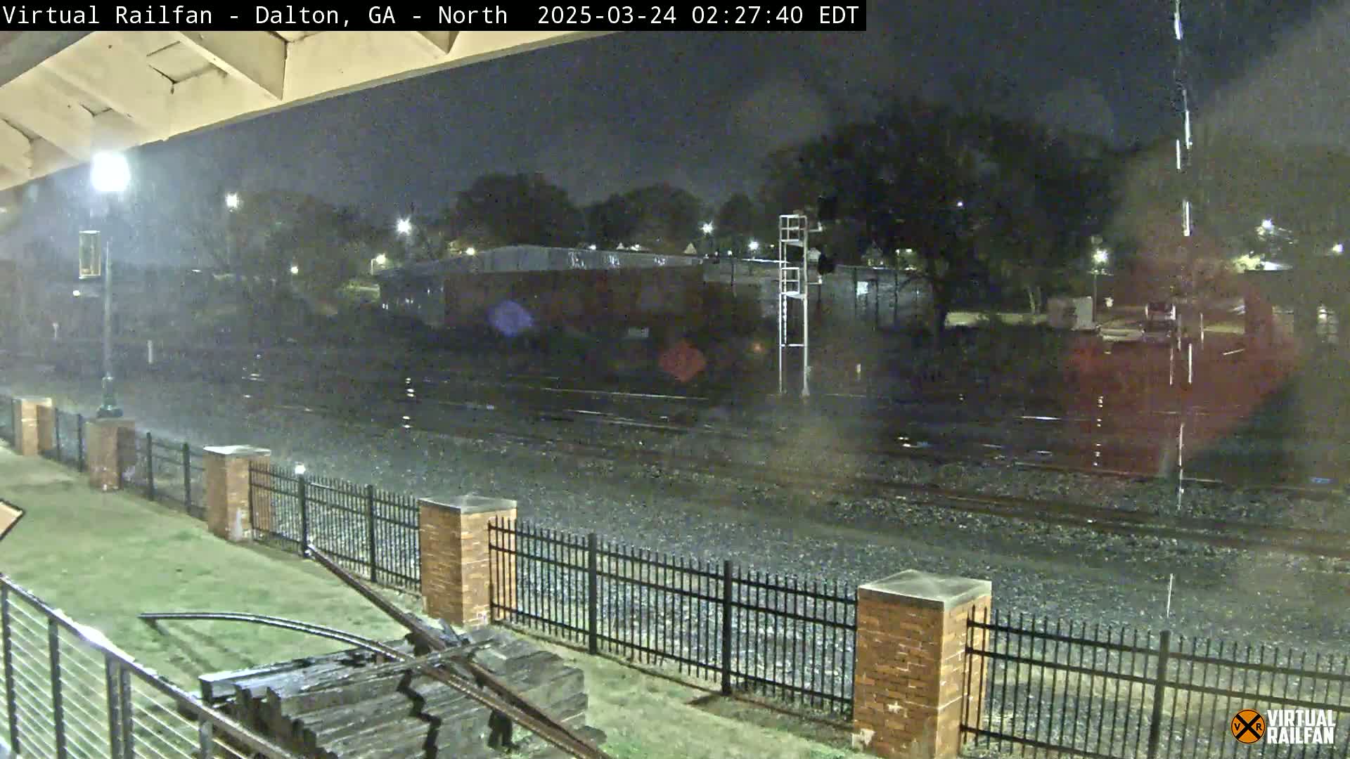 A nighttime view of rain falling on railroad tracks beside a brick fence, with a streetlight illuminating the area.