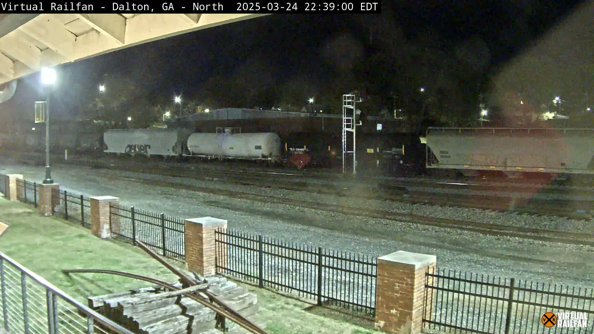 At night, a freight train sits on a rail line behind a brick fence and grassy area.
