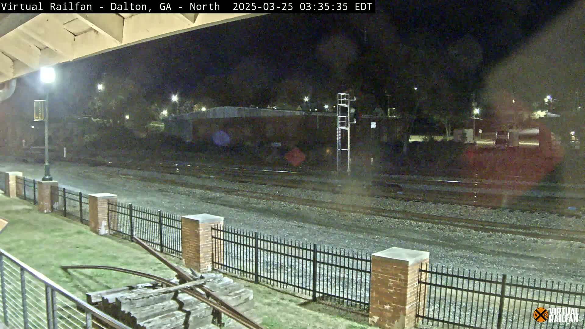 A nighttime view of railroad tracks, bordered by a black metal fence and brick pillars, with a grassy area and some old wooden railway equipment visible in the foreground.