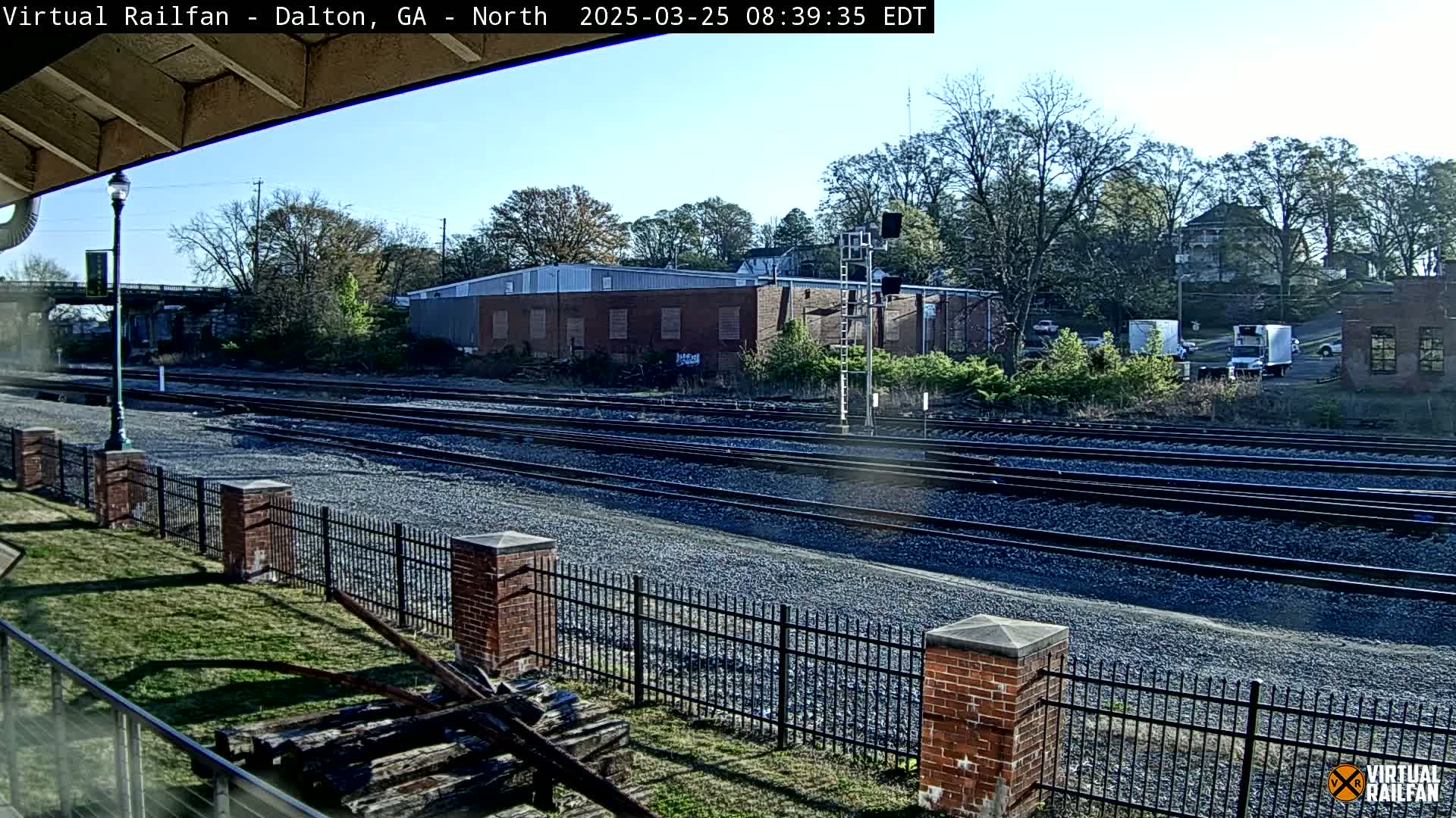 A sunny day reveals multiple train tracks bordered by a brick fence and sparse vegetation, with a brick building and trees in the background.