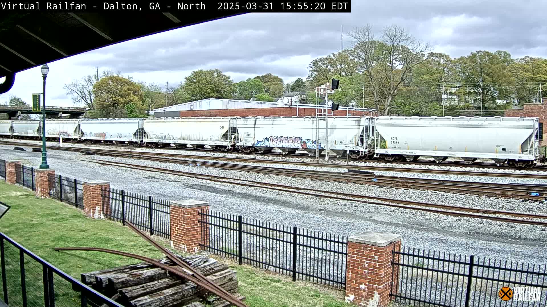 A long freight train of gray hopper cars, some with graffiti, sits on a railway track under a cloudy sky next to a grassy area with a brick fence.