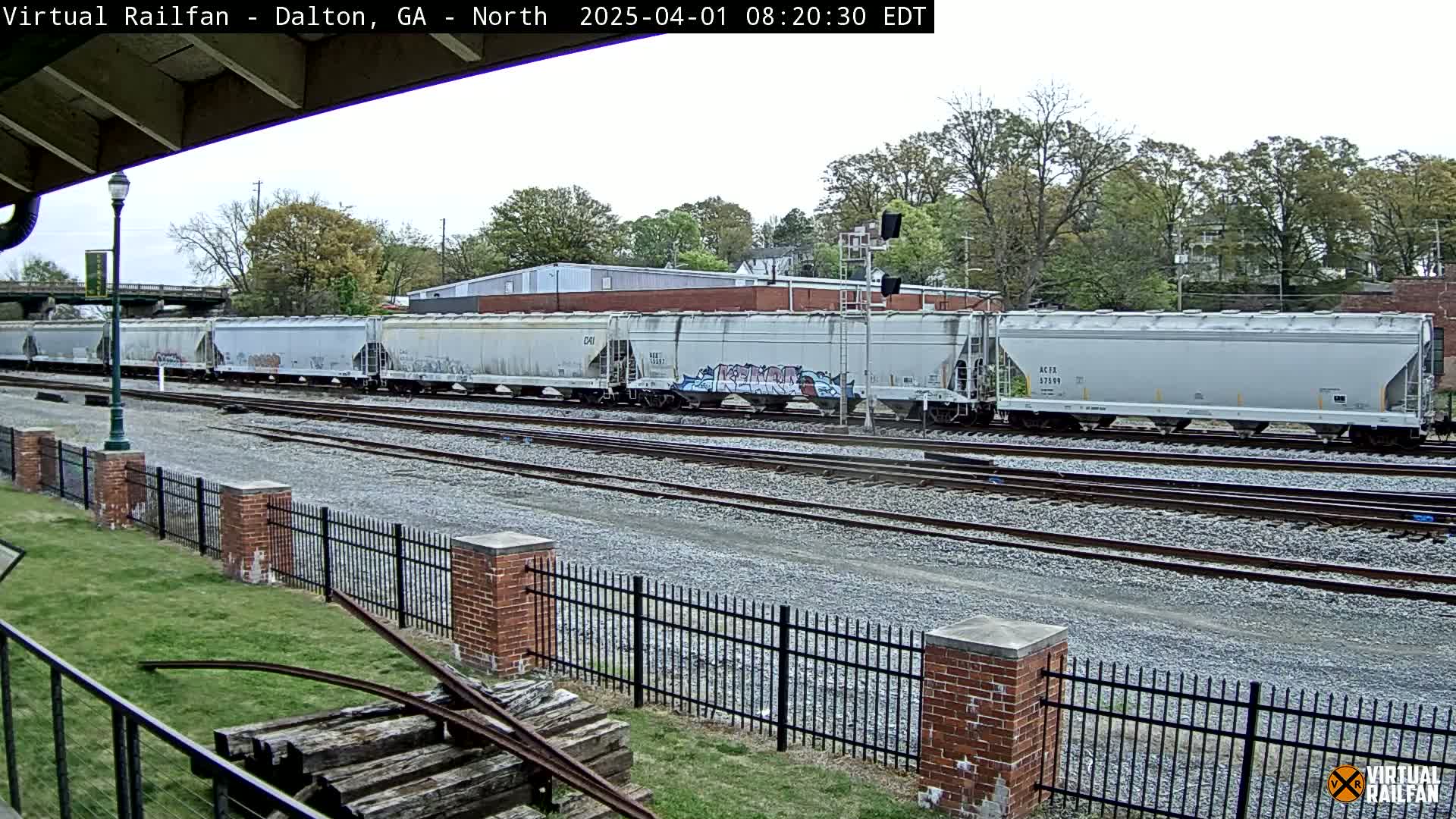 A long train of light gray covered hopper cars sits on a railway track under an overcast sky, beside a brick fence and grassy area.