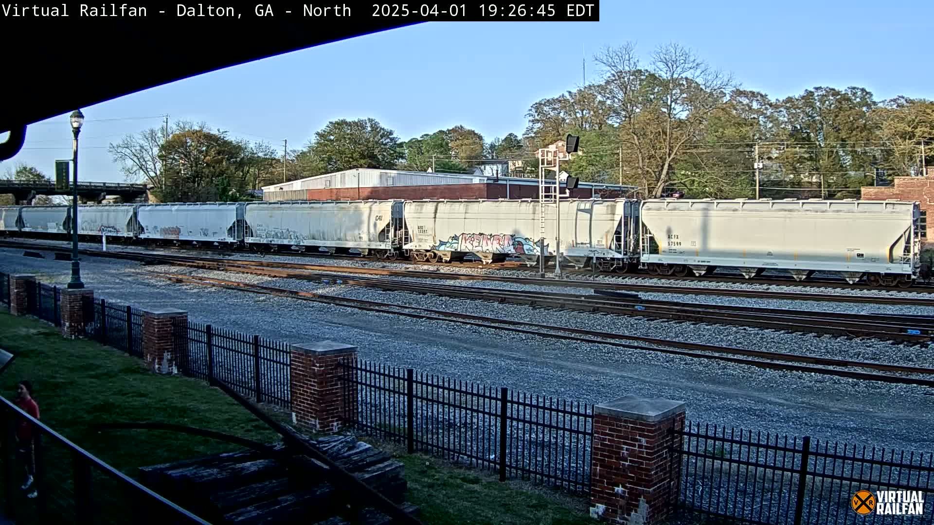 A long freight train of light gray covered hopper cars, some with graffiti, sits on railroad tracks under a clear, sunny sky.