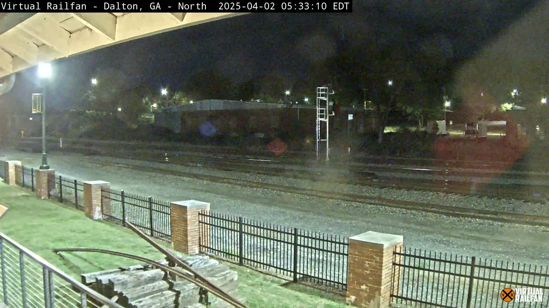 A nighttime view of railroad tracks, a brick fence, and a grassy area, with a tall metal structure in the background.