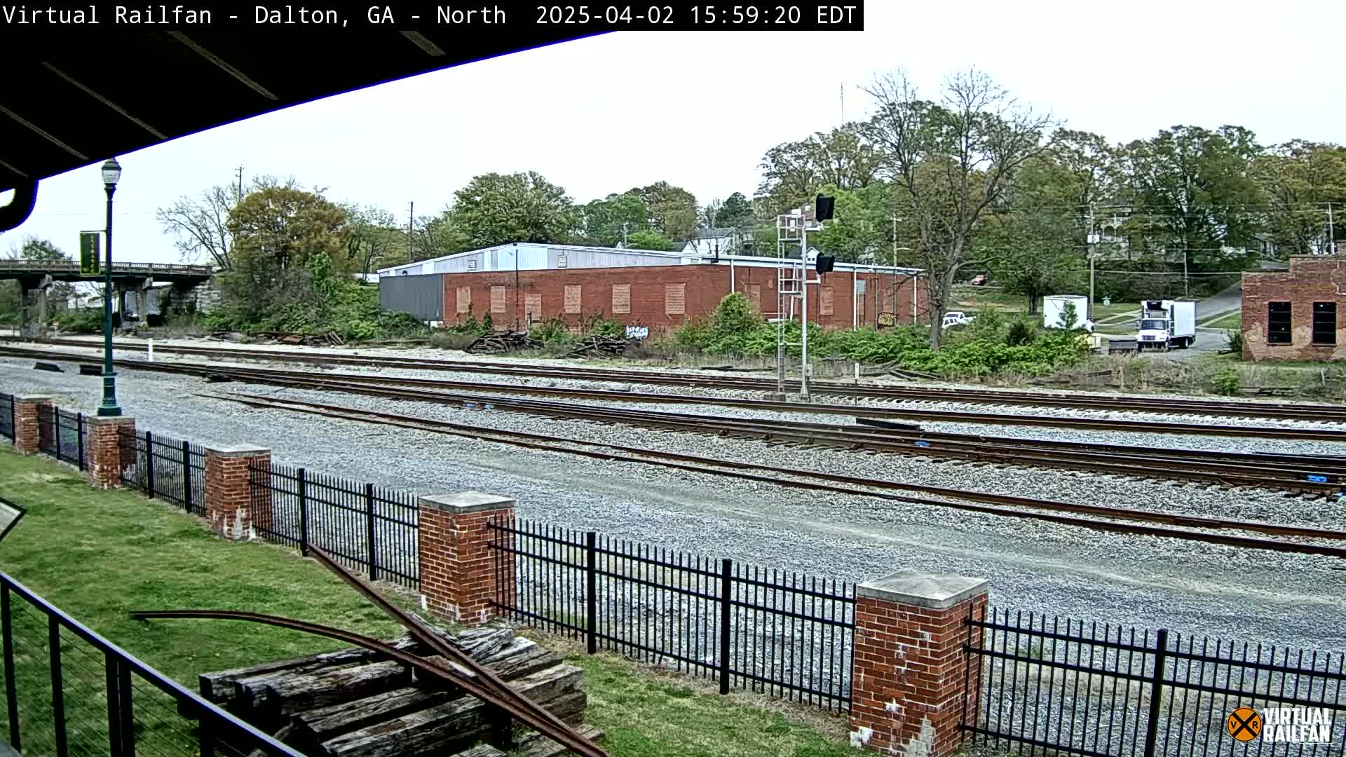 A view of multiple train tracks bordered by a brick fence on an overcast day, with buildings and trees in the background.
