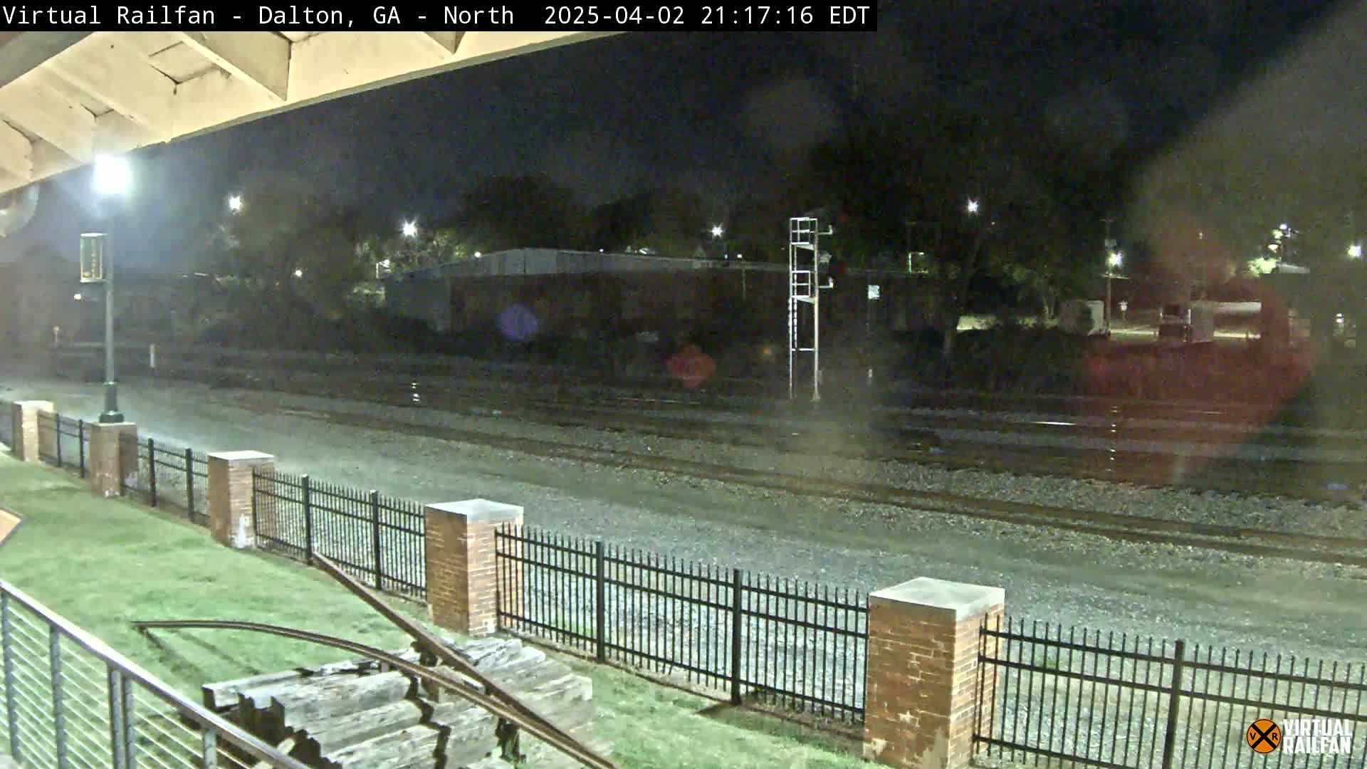 A nighttime view of railroad tracks beside a brick fence and grassy area, under a mostly clear sky.