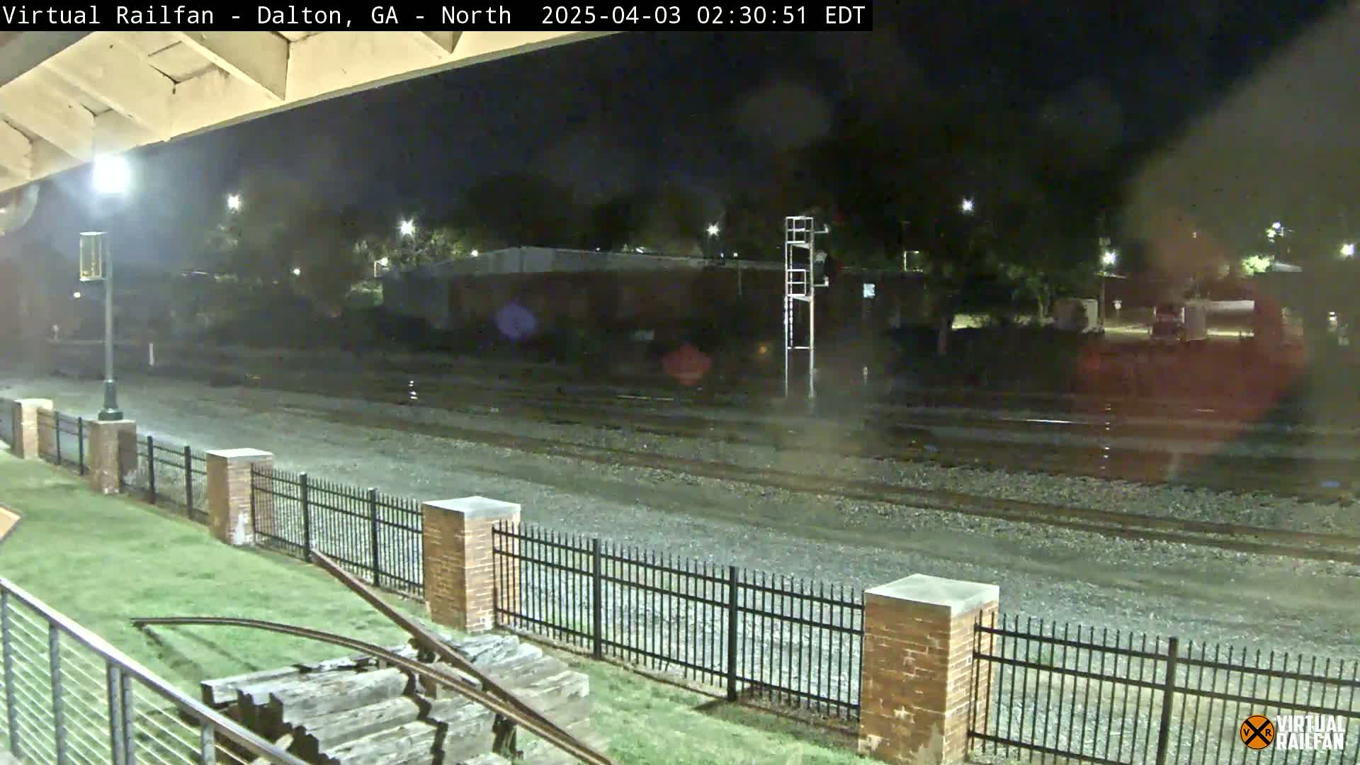 A nighttime view of wet railroad tracks behind a brick fence and grassy area, with a tall metal structure and some buildings visible in the background.