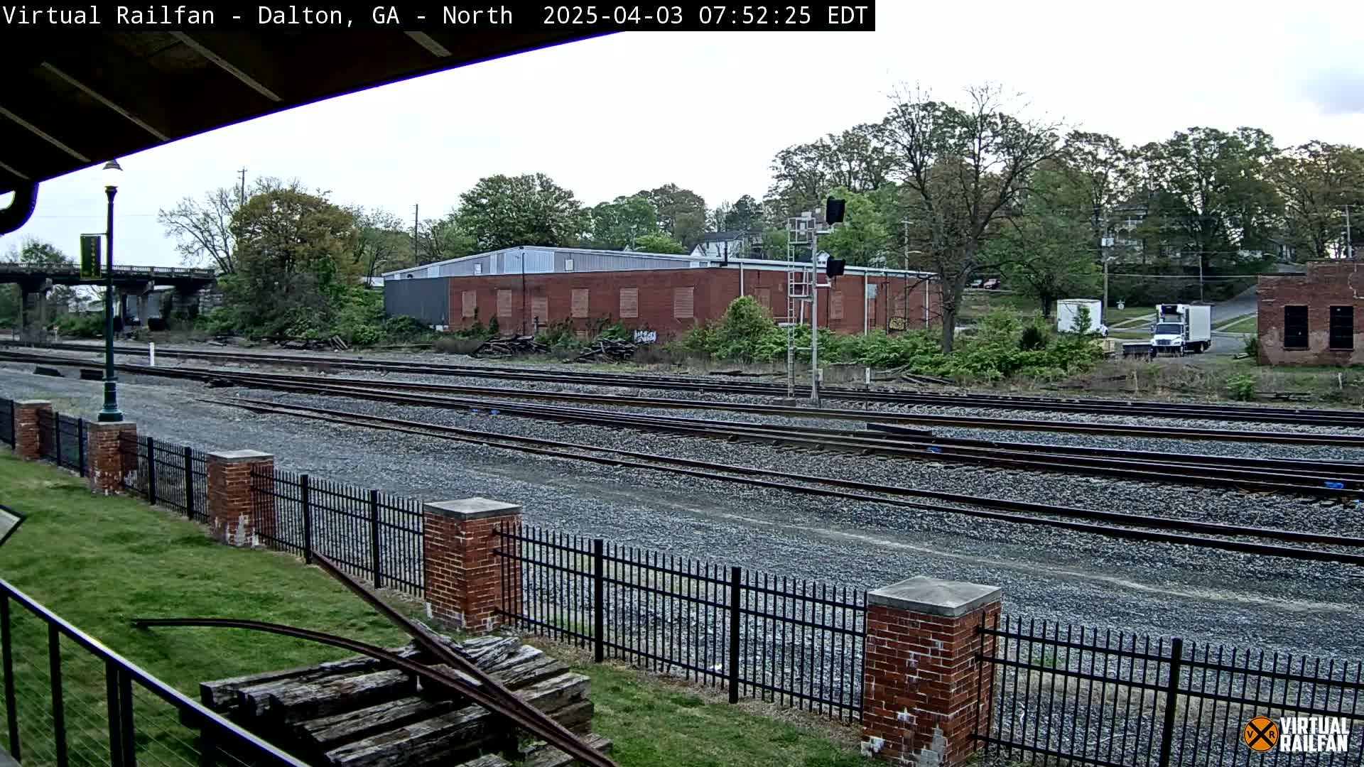 A view of several sets of train tracks beside a brick fence on an overcast day, with a brick building and trees in the background.