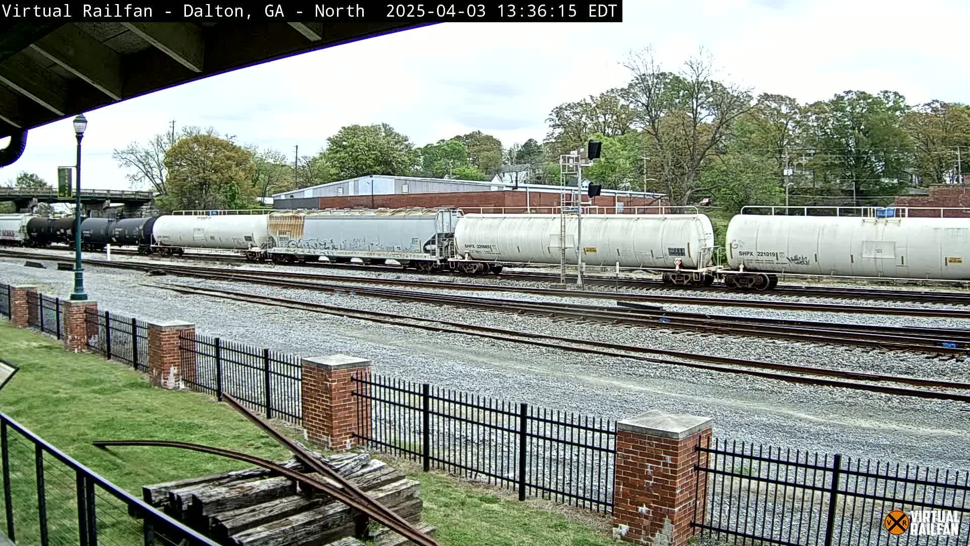 A long freight train with various tanker cars sits on a set of train tracks next to a brick fence on an overcast day.