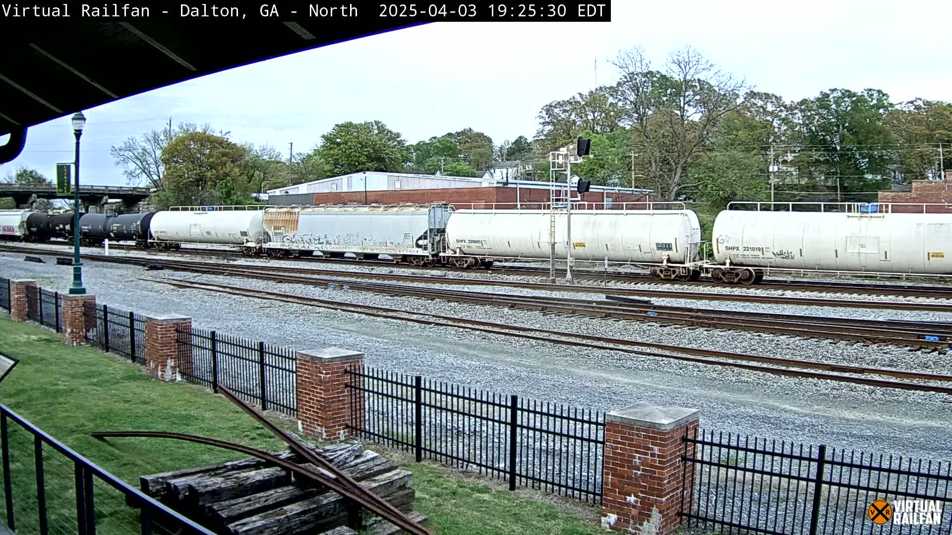 A long freight train of mostly white tanker cars sits on railroad tracks beside a brick fence and grassy area under a partly cloudy sky.