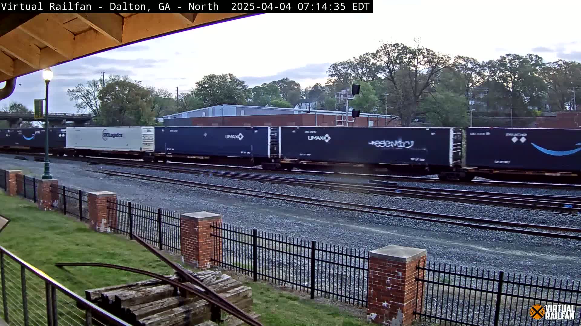 A freight train with various shipping containers passes by a brick fence on an overcast day.