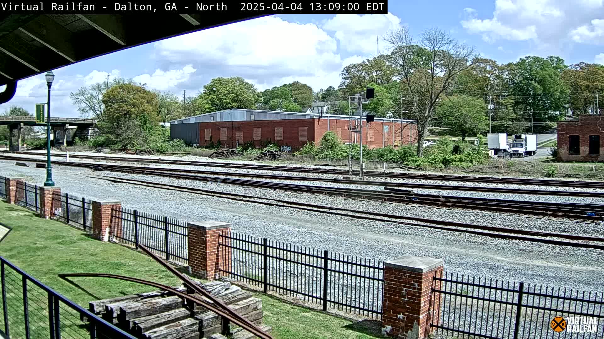 A partly cloudy day shows multiple train tracks extending into the distance, bordered by a brick fence and grassy area, with buildings and trees in the background.
