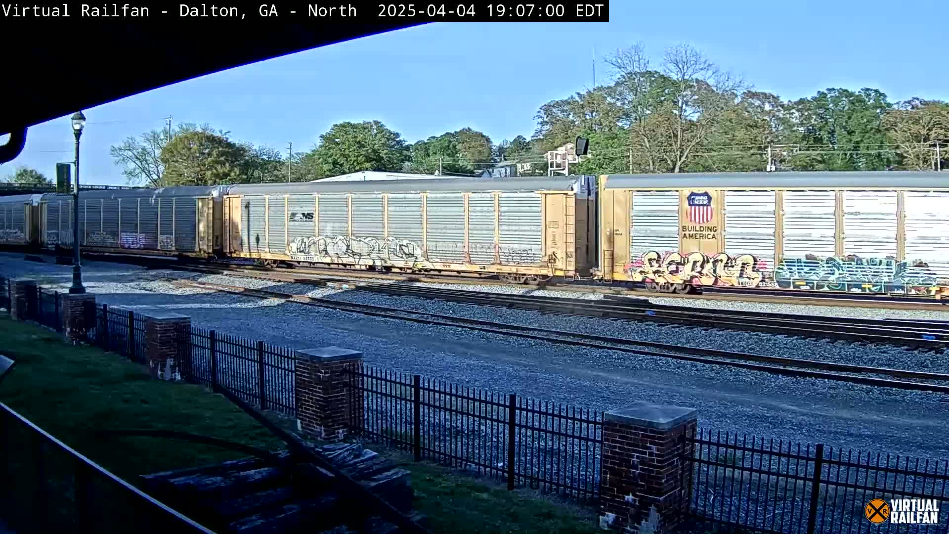 A long freight train with graffiti on some cars passes by under a mostly clear sky.