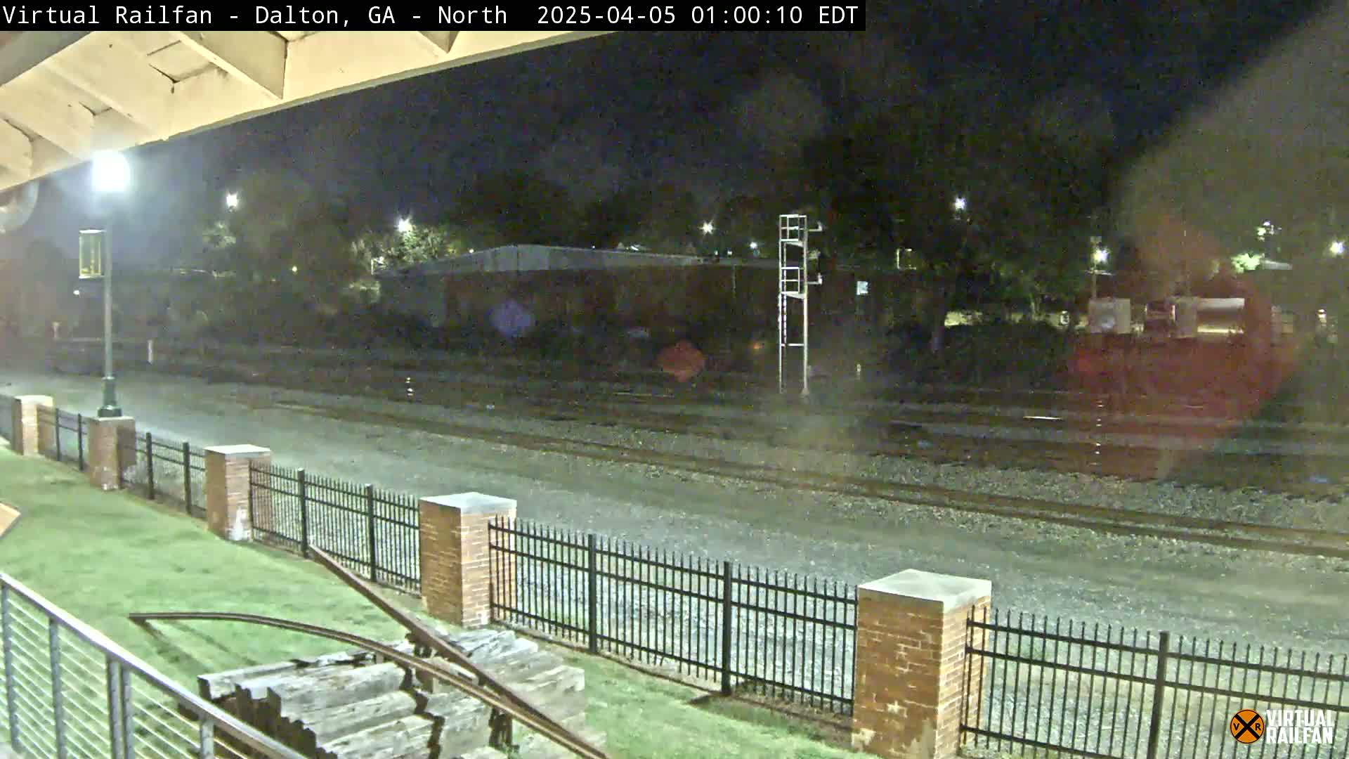 A nighttime view of railroad tracks, bordered by a brick fence, with a grassy area and some wooden structures visible in the foreground.