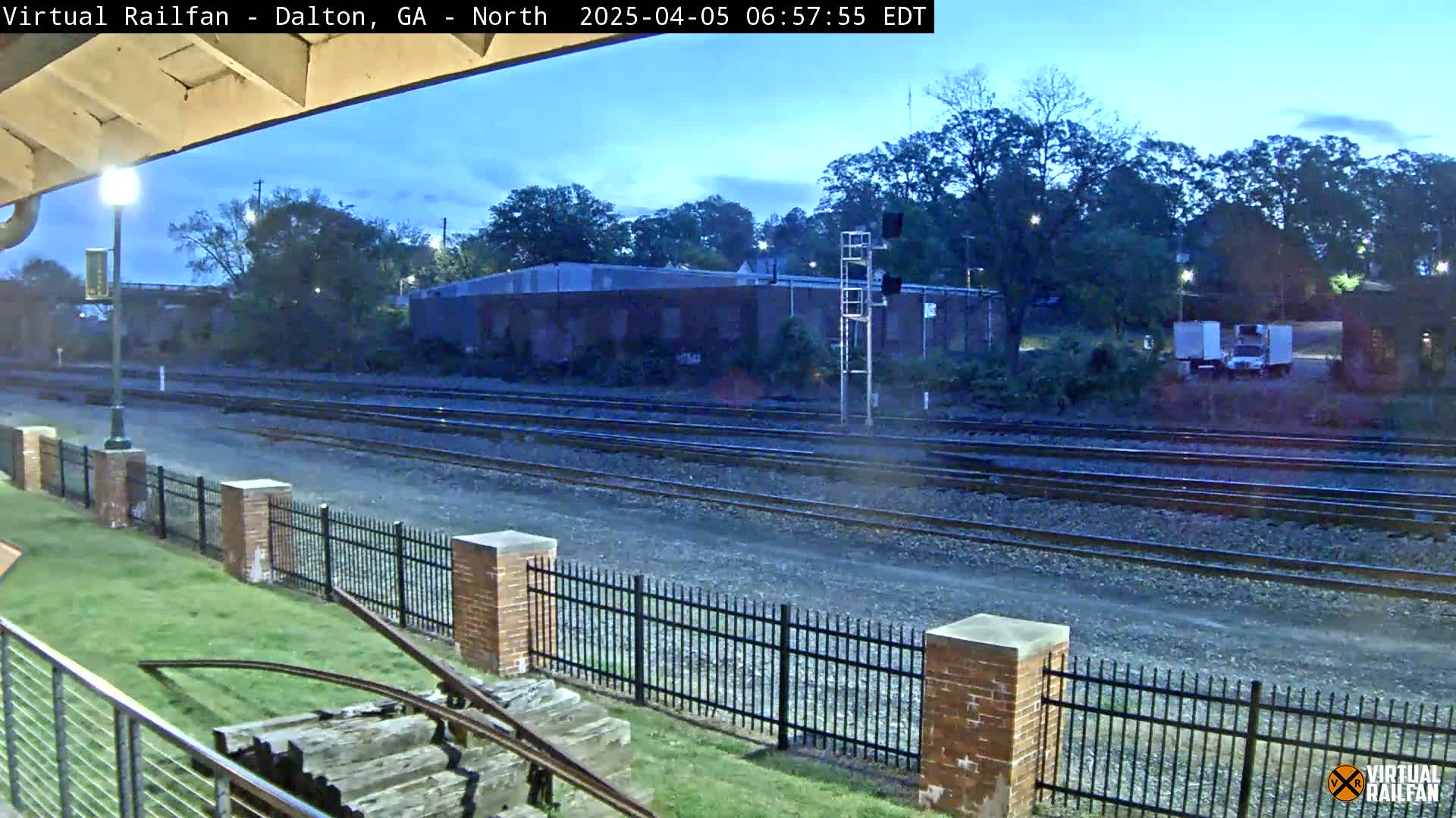 A view of train tracks at dawn, bordered by a brick fence and grassy area, with a few buildings and trees in the background.