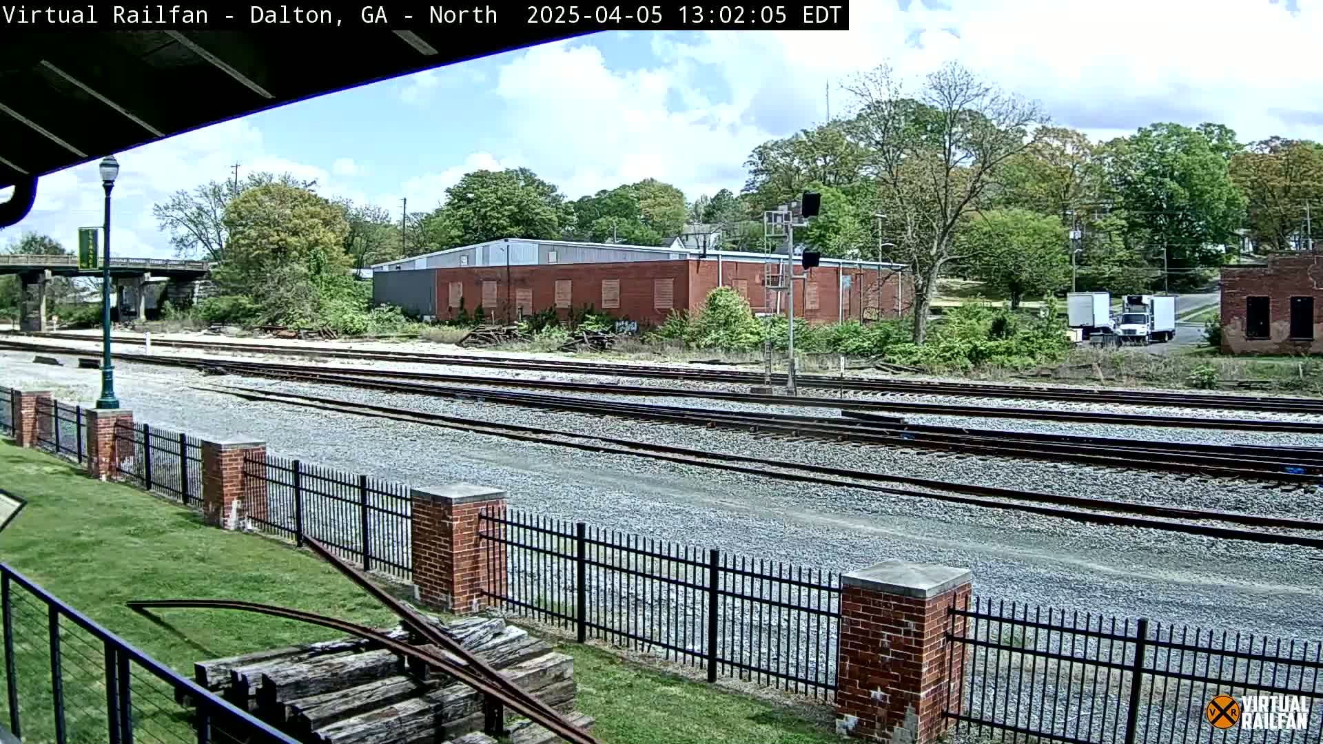 A partly cloudy day reveals a railroad yard with multiple train tracks, bordered by a brick fence and lush green grass, beyond which are industrial buildings and a line of trees.