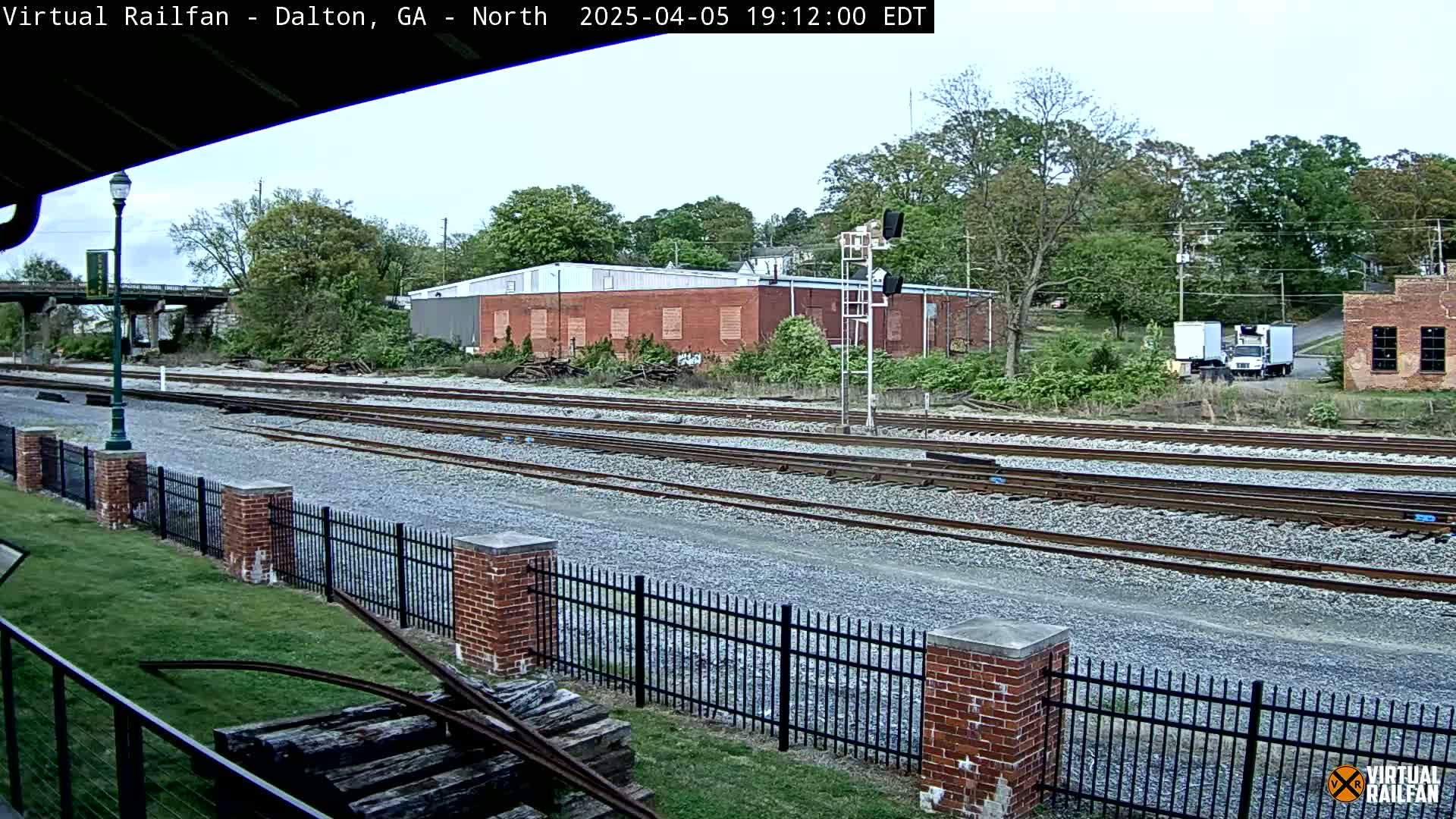 Multiple sets of train tracks extend into the distance alongside a brick fence, past a red brick building and several trees under a partly cloudy sky.