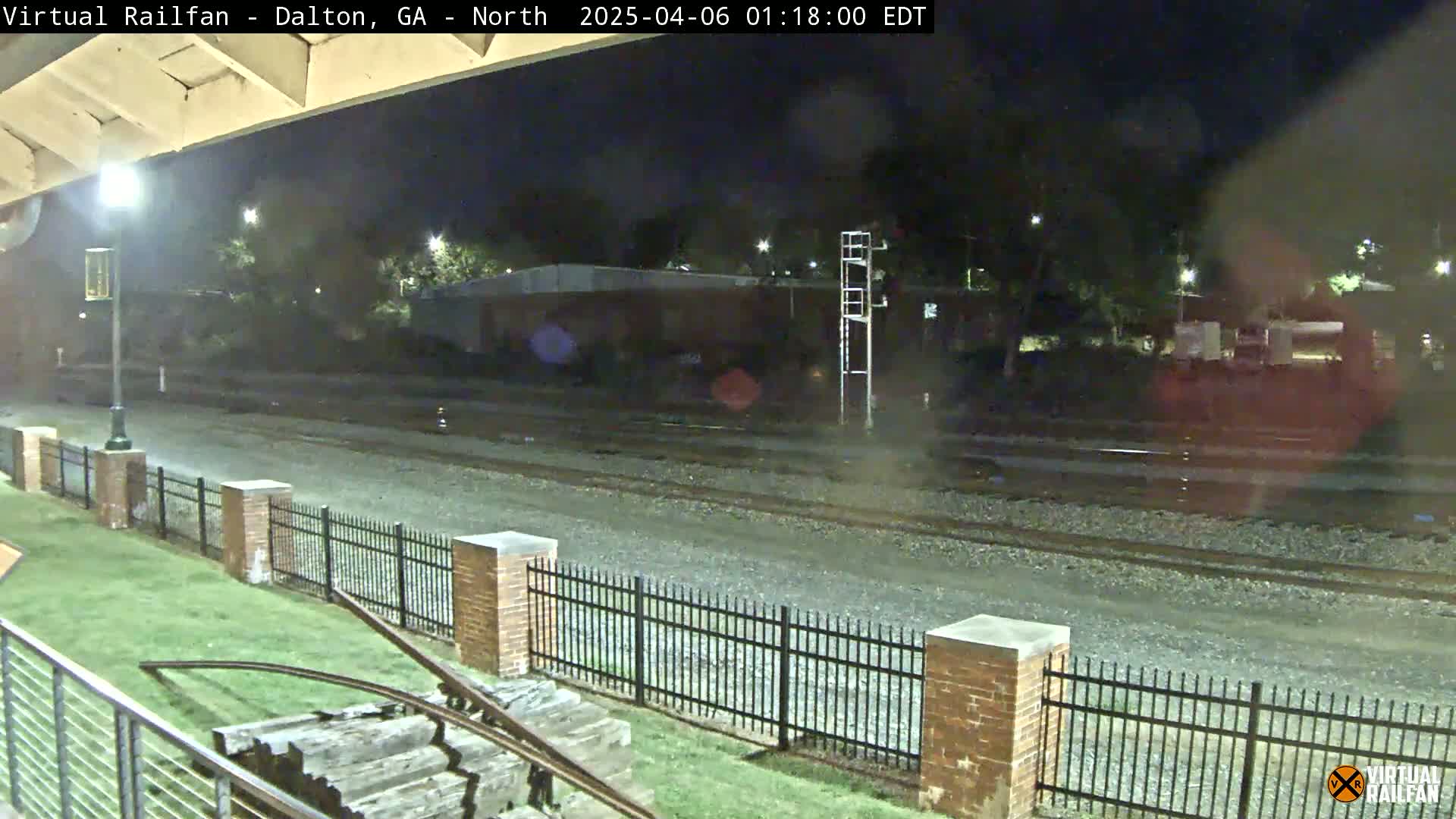 A nighttime view of railroad tracks bordered by a brick fence, with a grassy area and some wooden structures in the foreground.