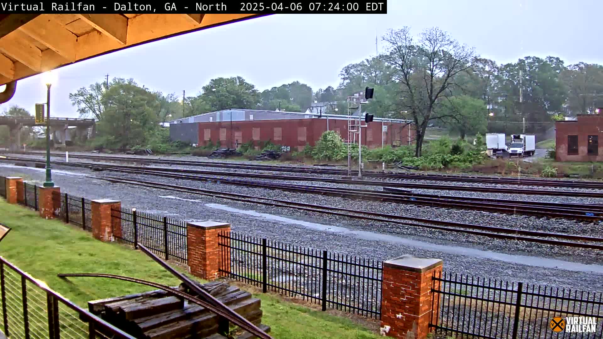 Rain falls on a railroad yard bordered by a brick fence and lush green grass, with industrial buildings and a few trucks visible in the background.