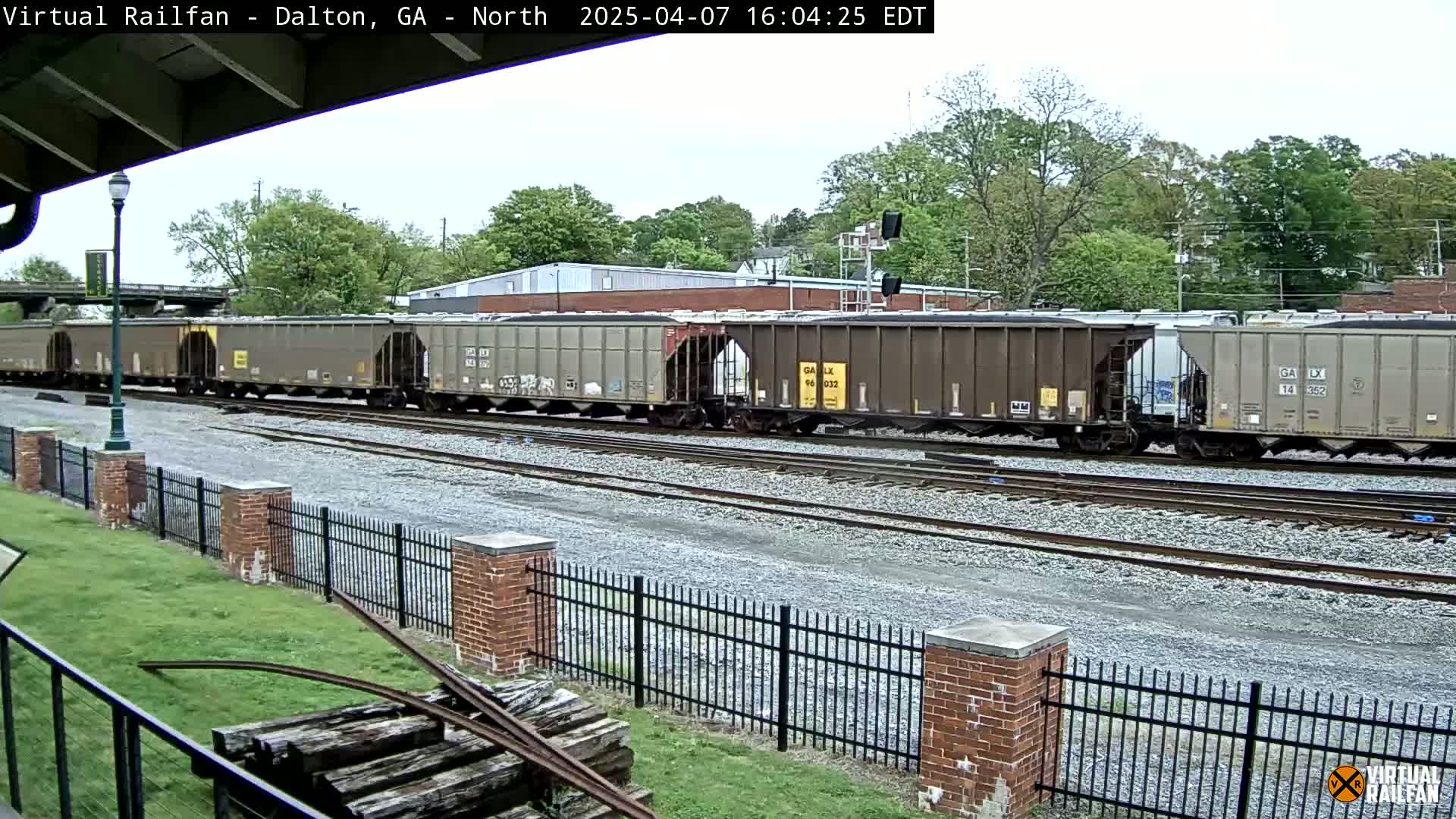 A long freight train of covered hopper cars sits on a railway track under a partly cloudy sky, next to a brick fence and grassy area.