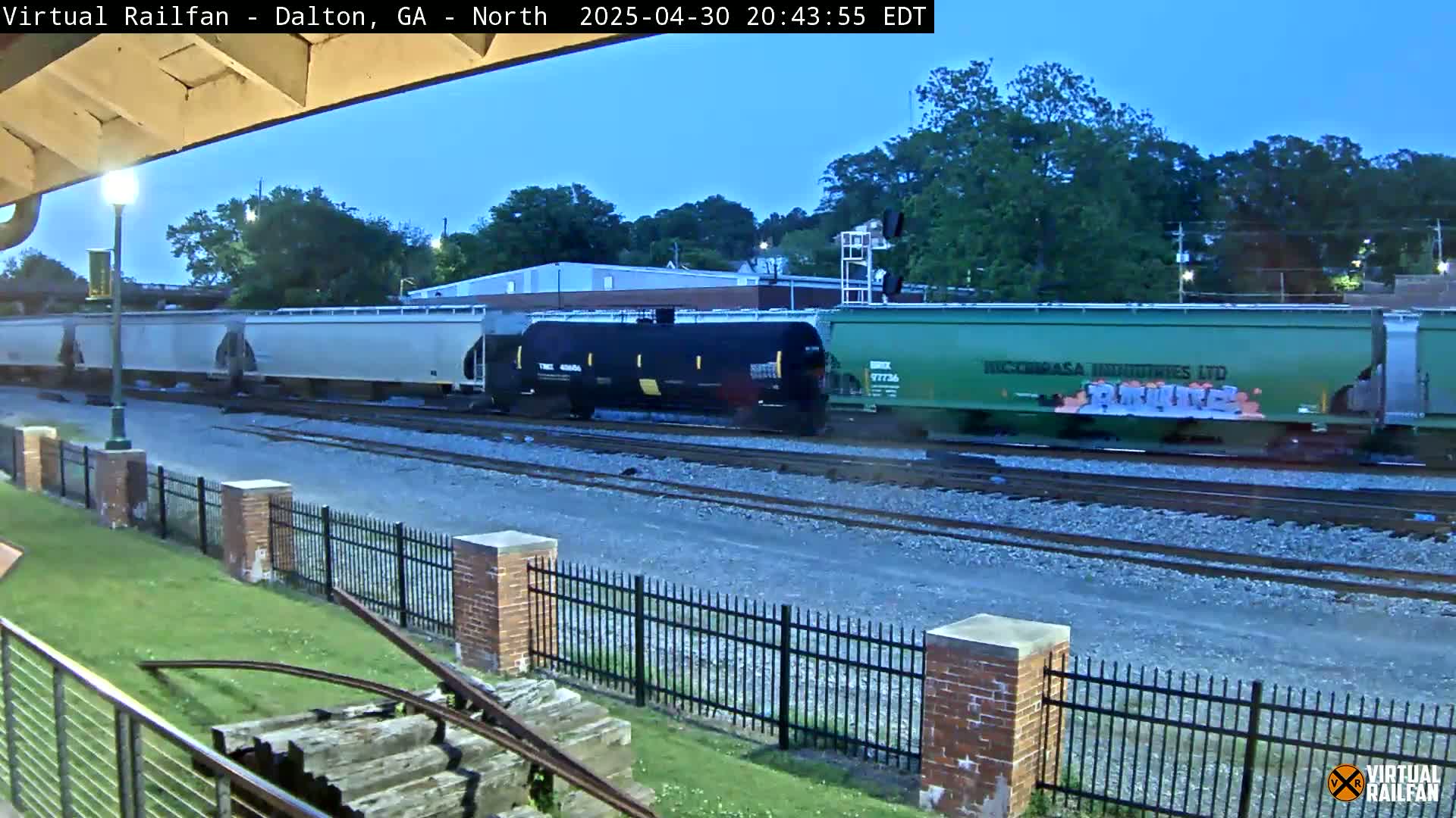 A freight train with various colored cars sits on railway tracks under a twilight sky next to a brick fence and grassy area.
