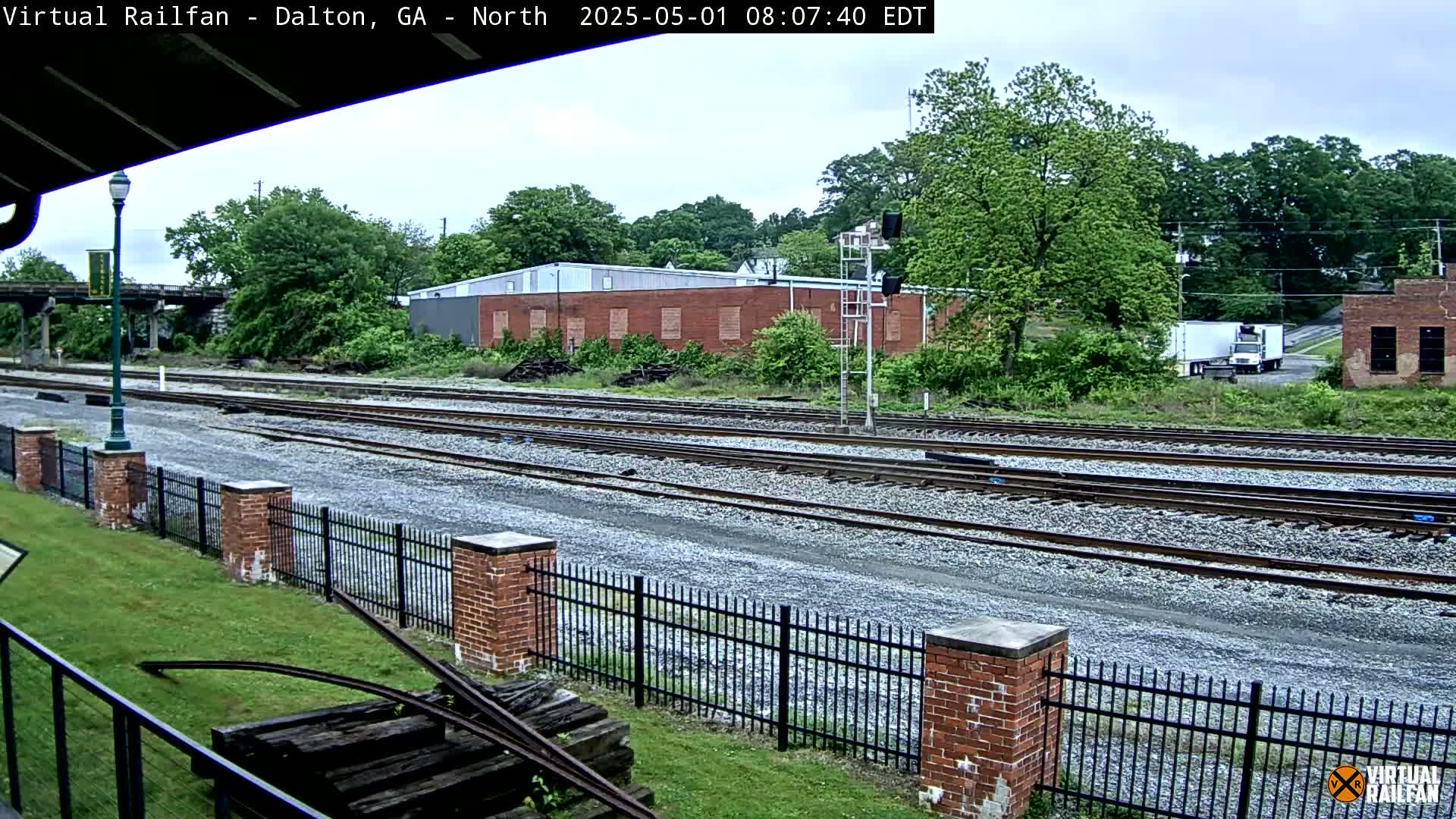 Multiple train tracks extend into the distance, bordered by a brick fence and lush greenery under an overcast sky.