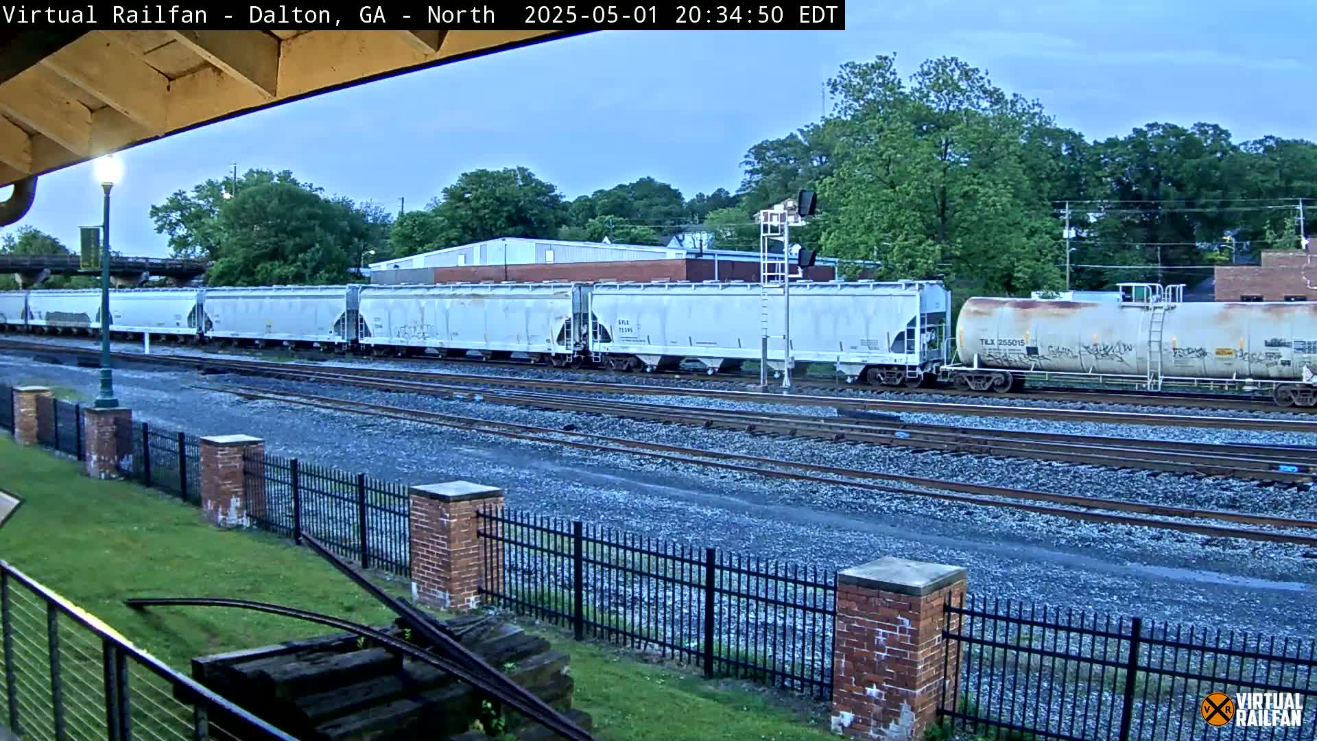 A long freight train sits on railway tracks next to a brick fence under a partly cloudy evening sky.