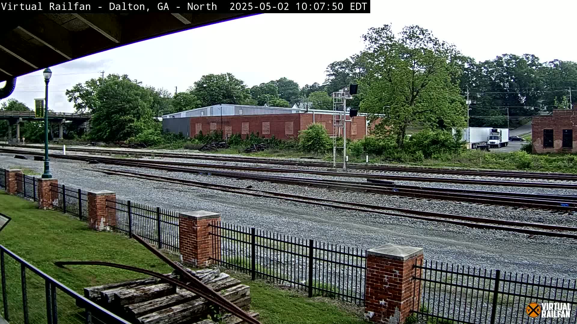 Multiple sets of train tracks extend into the distance, bordered by a brick fence and lush green vegetation under an overcast sky.