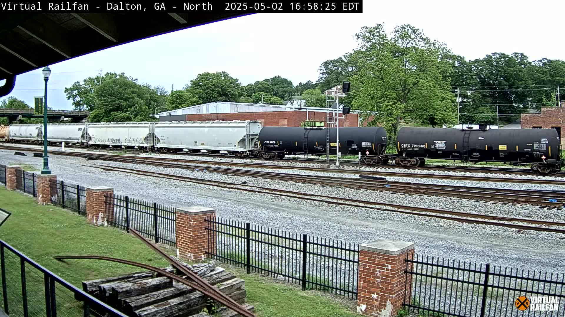 A freight train consisting of several light gray covered hopper cars and dark-colored tanker cars sits on a set of railroad tracks on an overcast day, beside a brick fence and lush green grass.