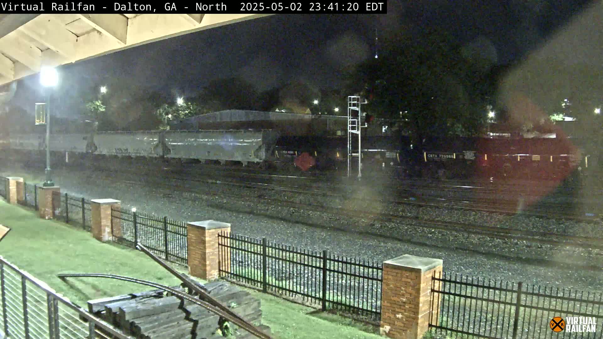 A nighttime view of a freight train passing by a fenced-in area, with dark skies and possibly light rain.