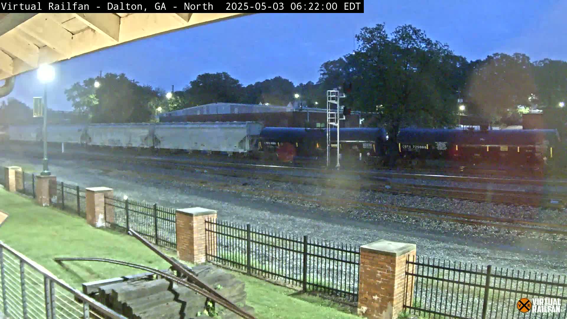A freight train with gray and black cars passes by under a twilight sky next to a grassy area enclosed by a brick fence.