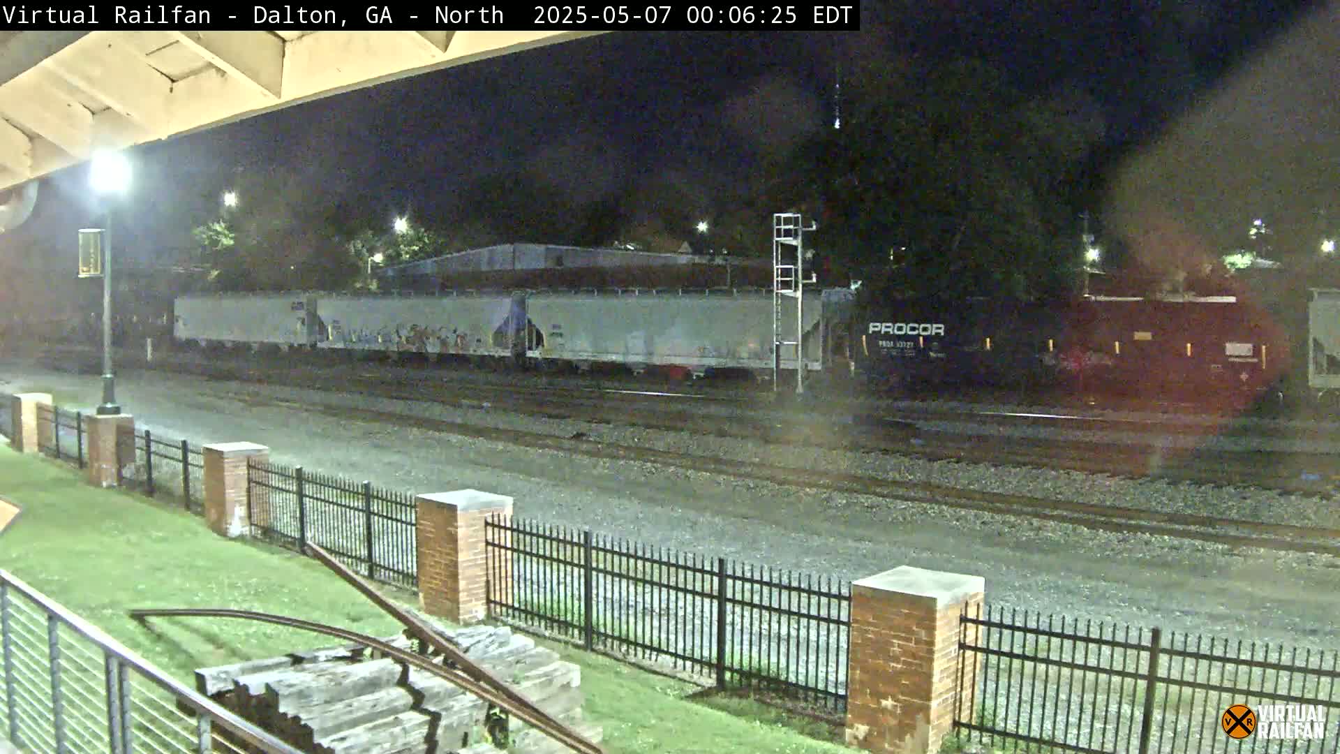 At night, a freight train passes by a brick fence and grassy area next to a railway track.
