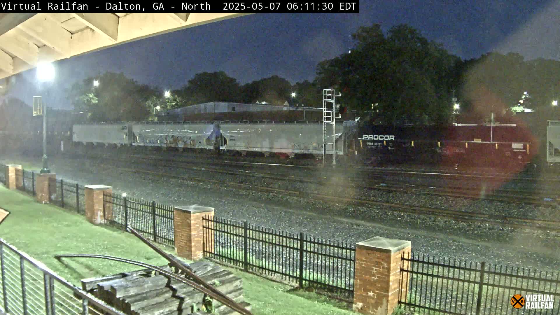 A freight train passes by under nighttime, rainy conditions, viewed from across a black metal fence.