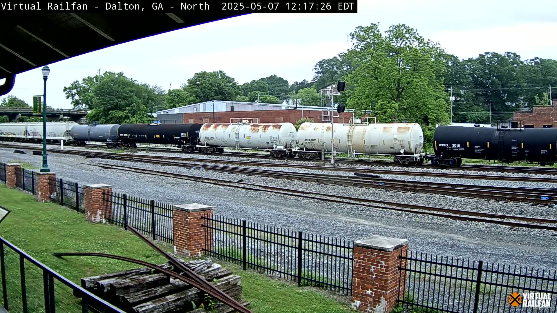 A long freight train of tanker cars sits on a railway track next to a brick fence and grassy area under an overcast sky.