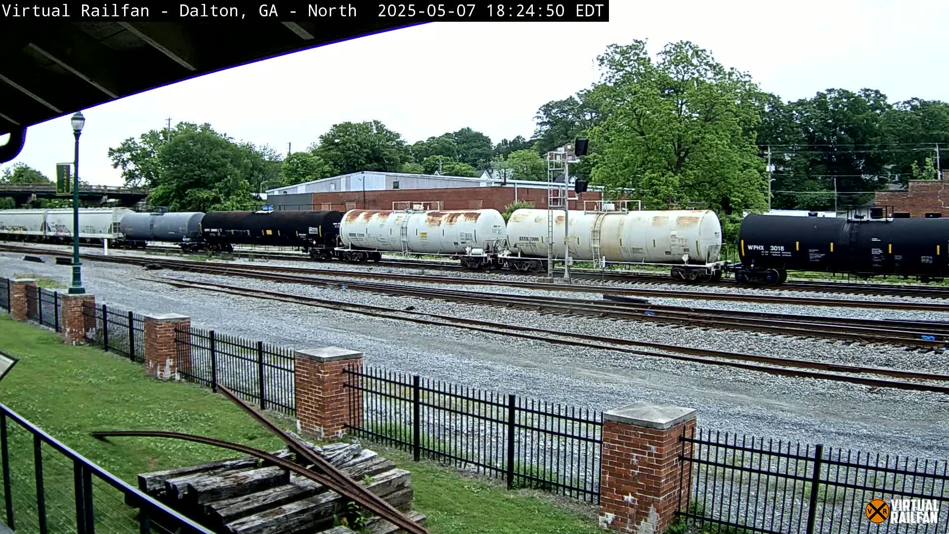 A long freight train of various tanker cars sits on a railway track under an overcast sky next to a brick fence and grassy area.