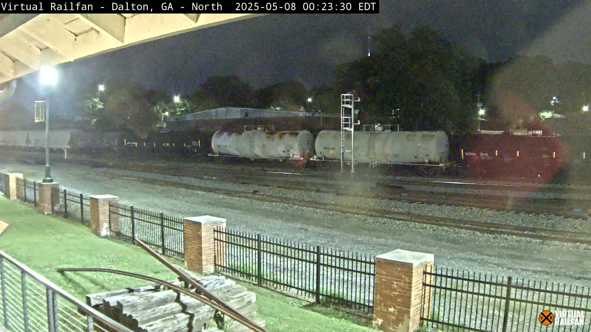 At night, several train cars sit on railway tracks next to a brick fence and grassy area.