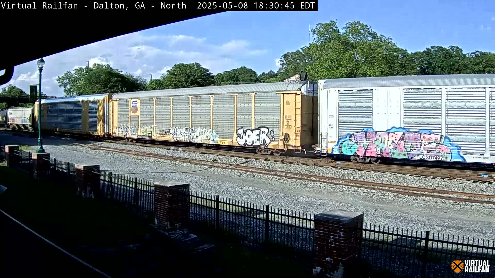 Under a partly cloudy sky, a freight train with graffiti-covered boxcars sits on a railway track next to a chain-link fence.
