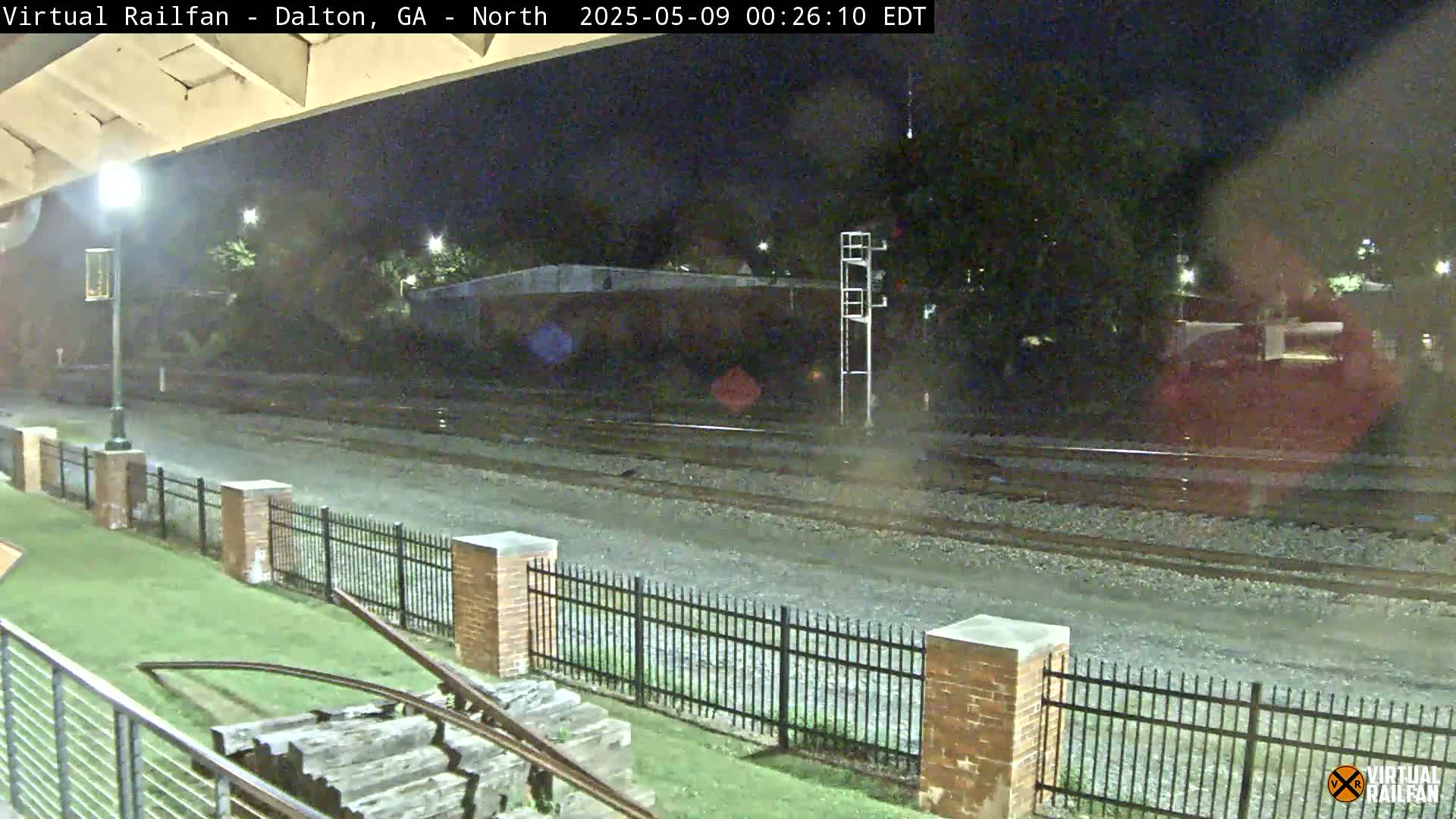 A nighttime view of railroad tracks and a brick fence, with some vegetation and a lamppost visible under a clear sky.