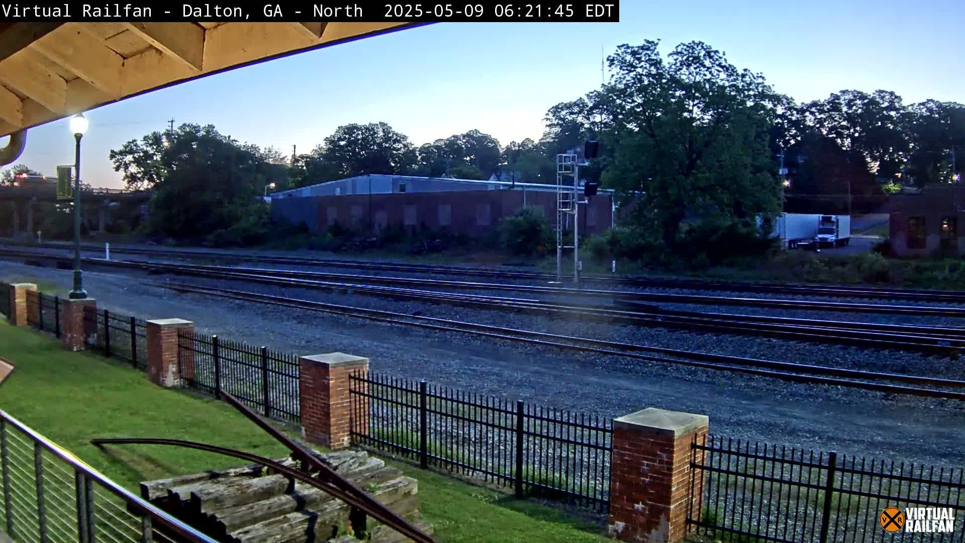 A view of multiple train tracks at dusk, bordered by a brick fence and lush greenery, with a large industrial building in the background.
