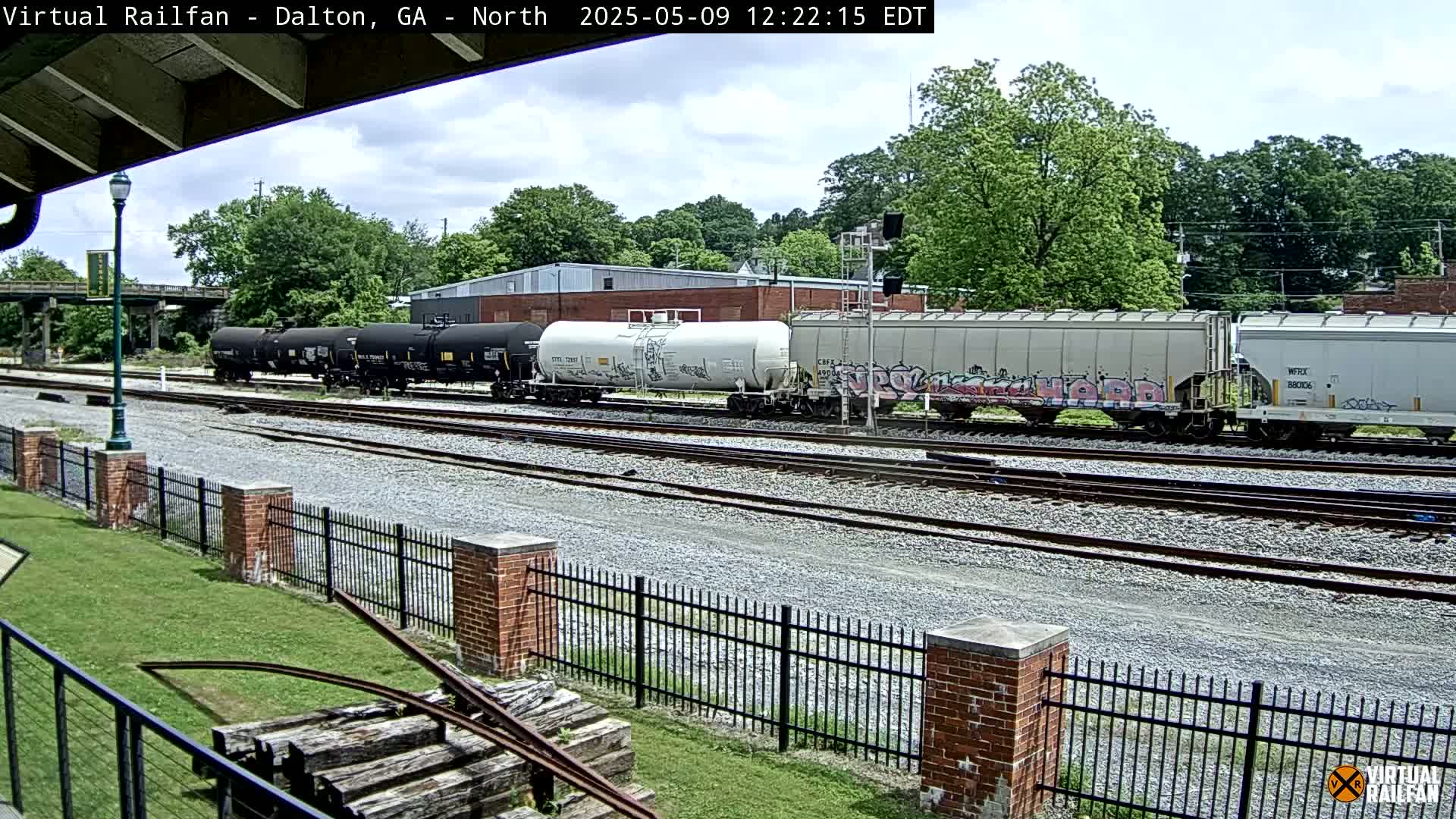 Several freight cars sit on railroad tracks next to a brick fence on a partly sunny day.