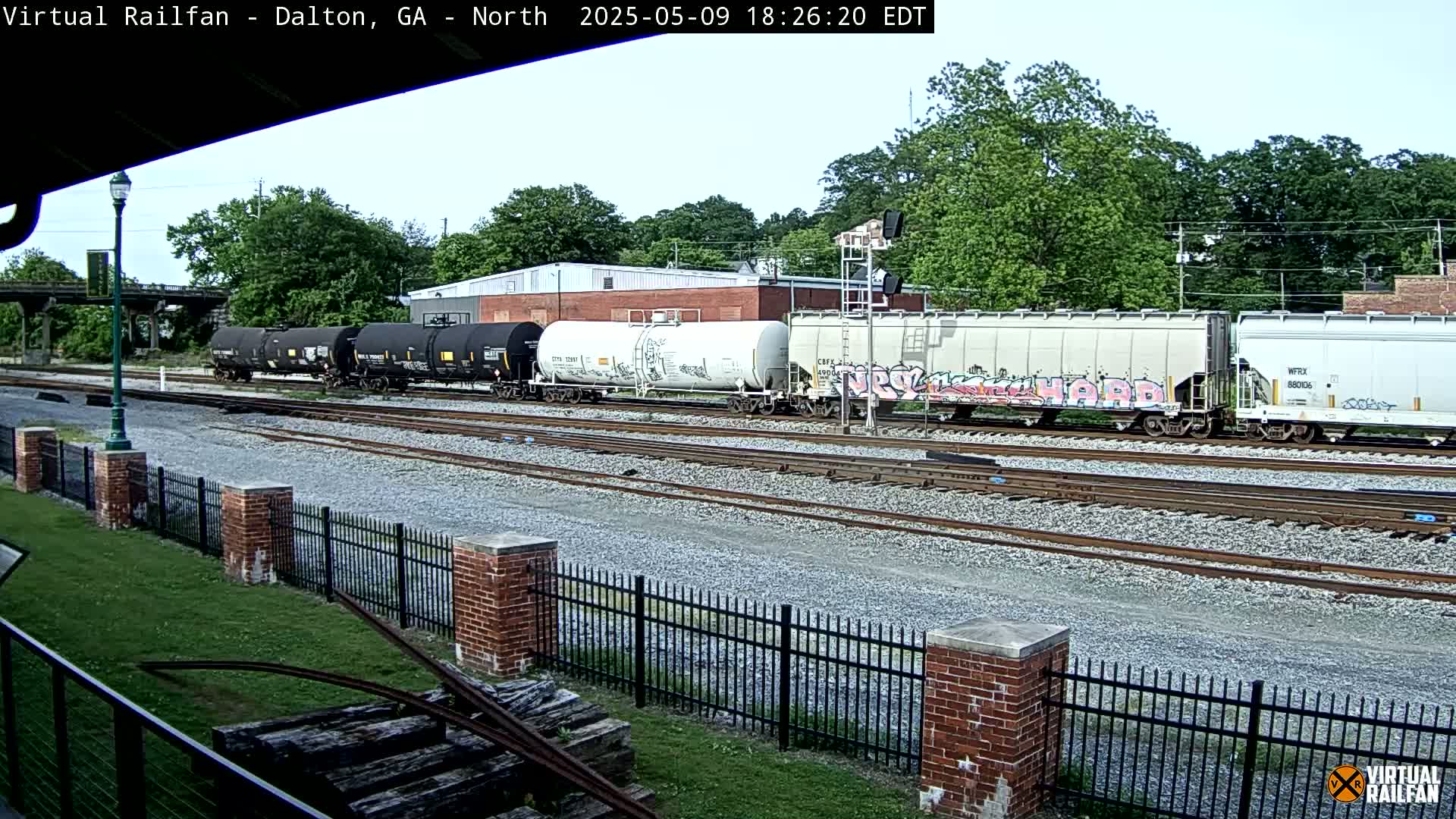 A long freight train with graffiti on some cars sits on a set of tracks on a partly sunny day, next to a brick fence and grassy area.