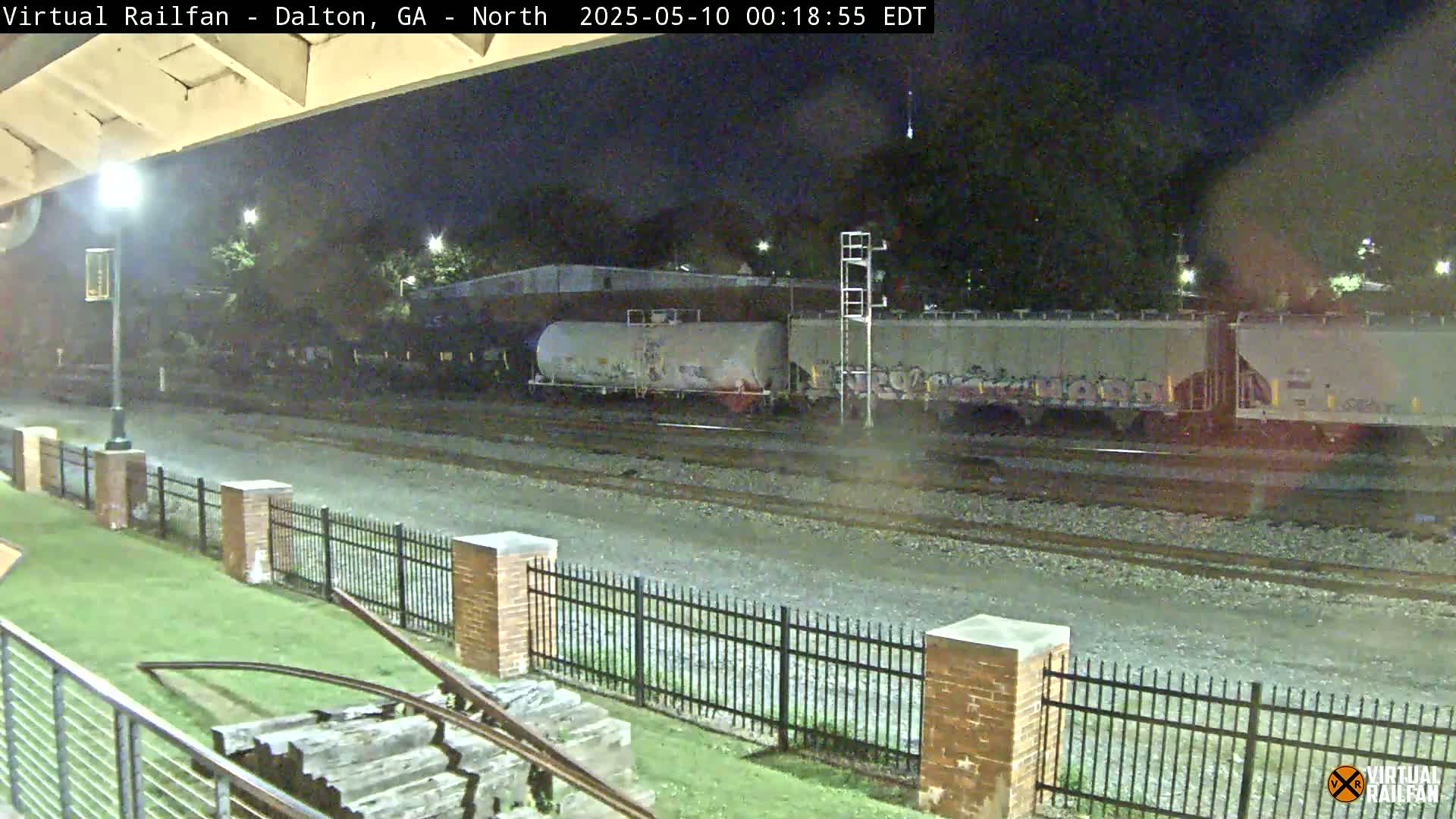 At night, a freight train sits on railroad tracks next to a brick fence and grassy area.