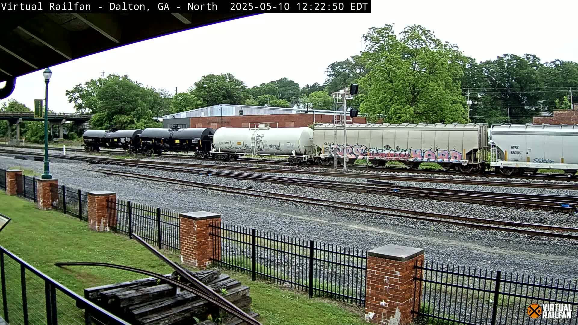 A freight train with various cars, some with graffiti, sits on a rail line next to a brick fence on an overcast day.