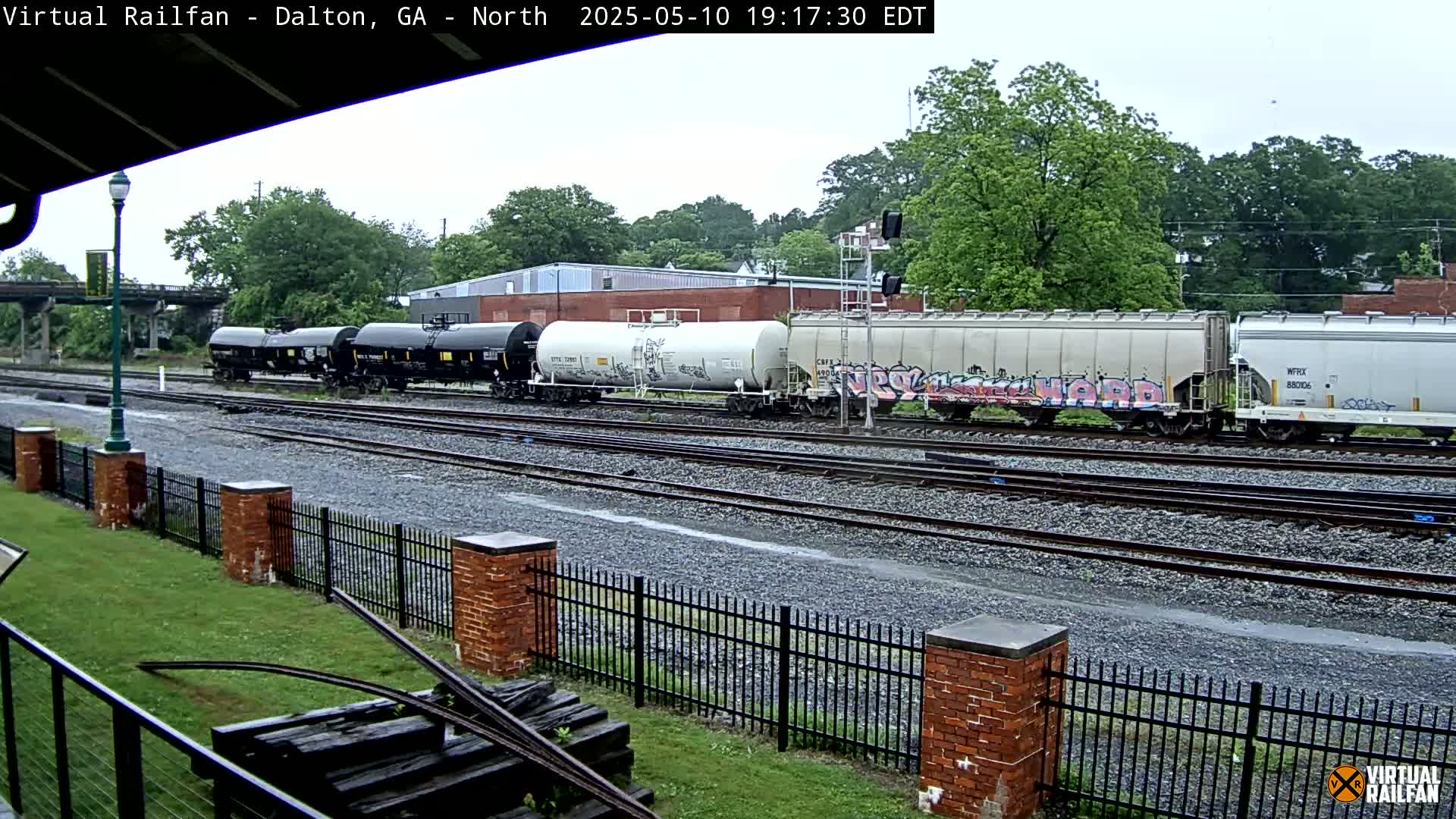 A freight train with graffiti-covered cars sits on a set of railway tracks in the rain, behind a short brick fence and lush green grass.