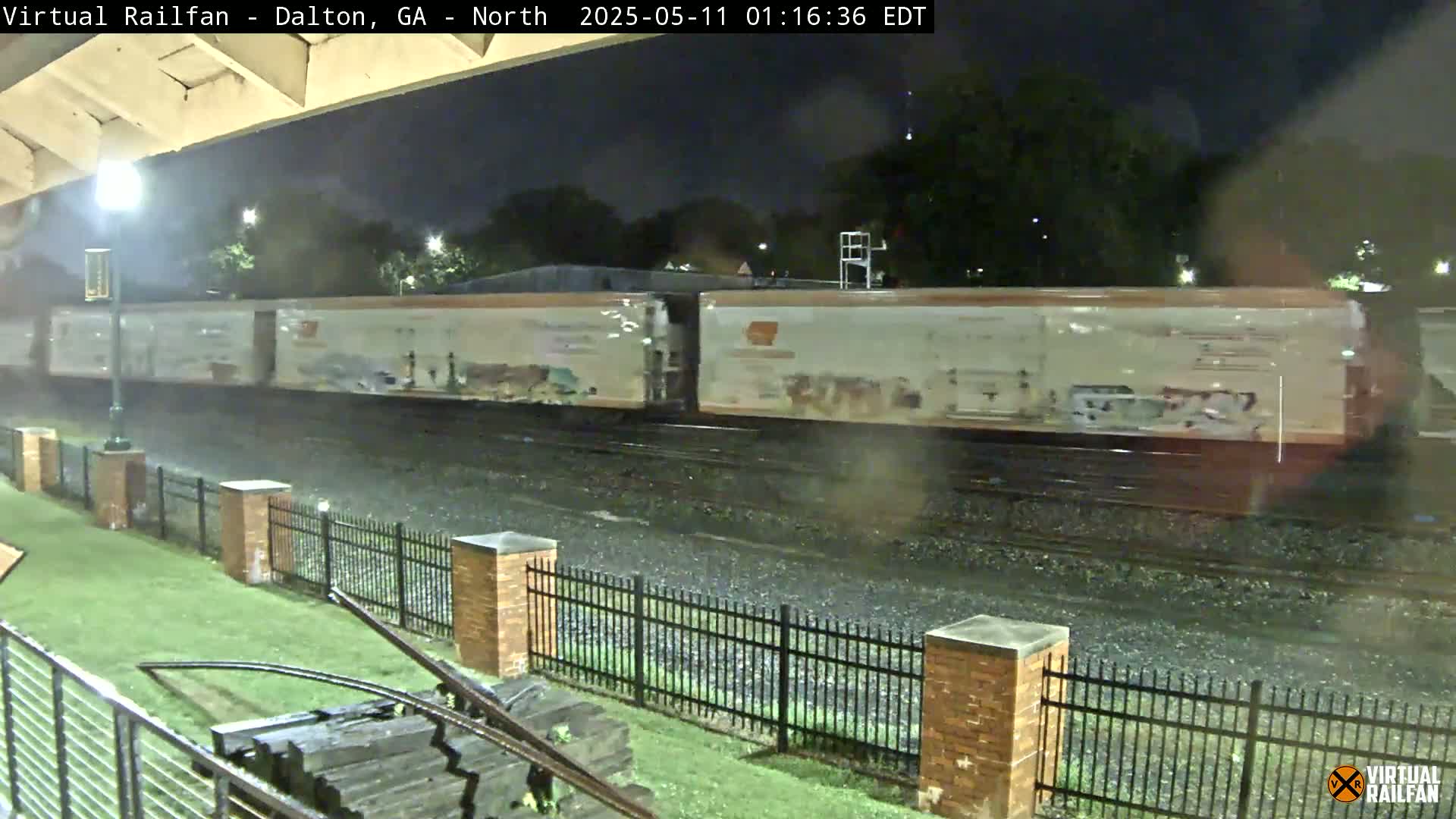 At night, under a light rain, a long freight train passes by a black metal fence and a grassy area.