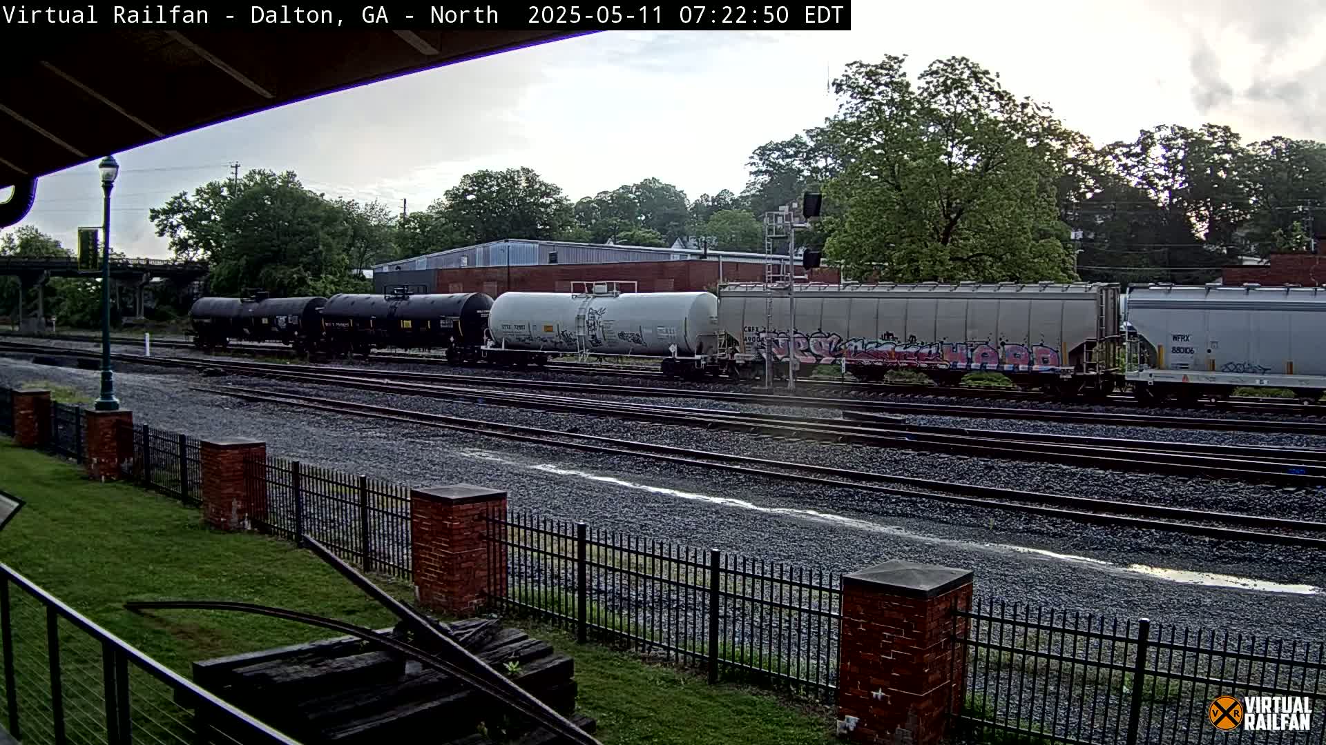 A freight train sits on a railway track under an overcast sky, next to a brick fence and grassy area.